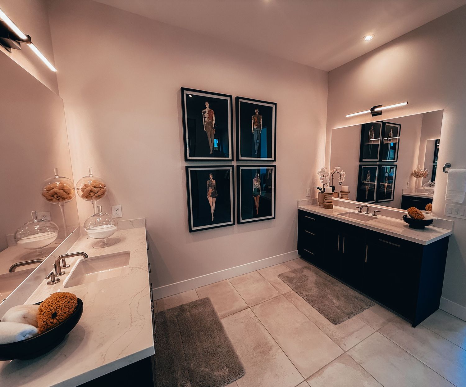 Elegant bathroom with dual vanities, marble countertops, large mirror, and four framed fashion art prints on pink walls.