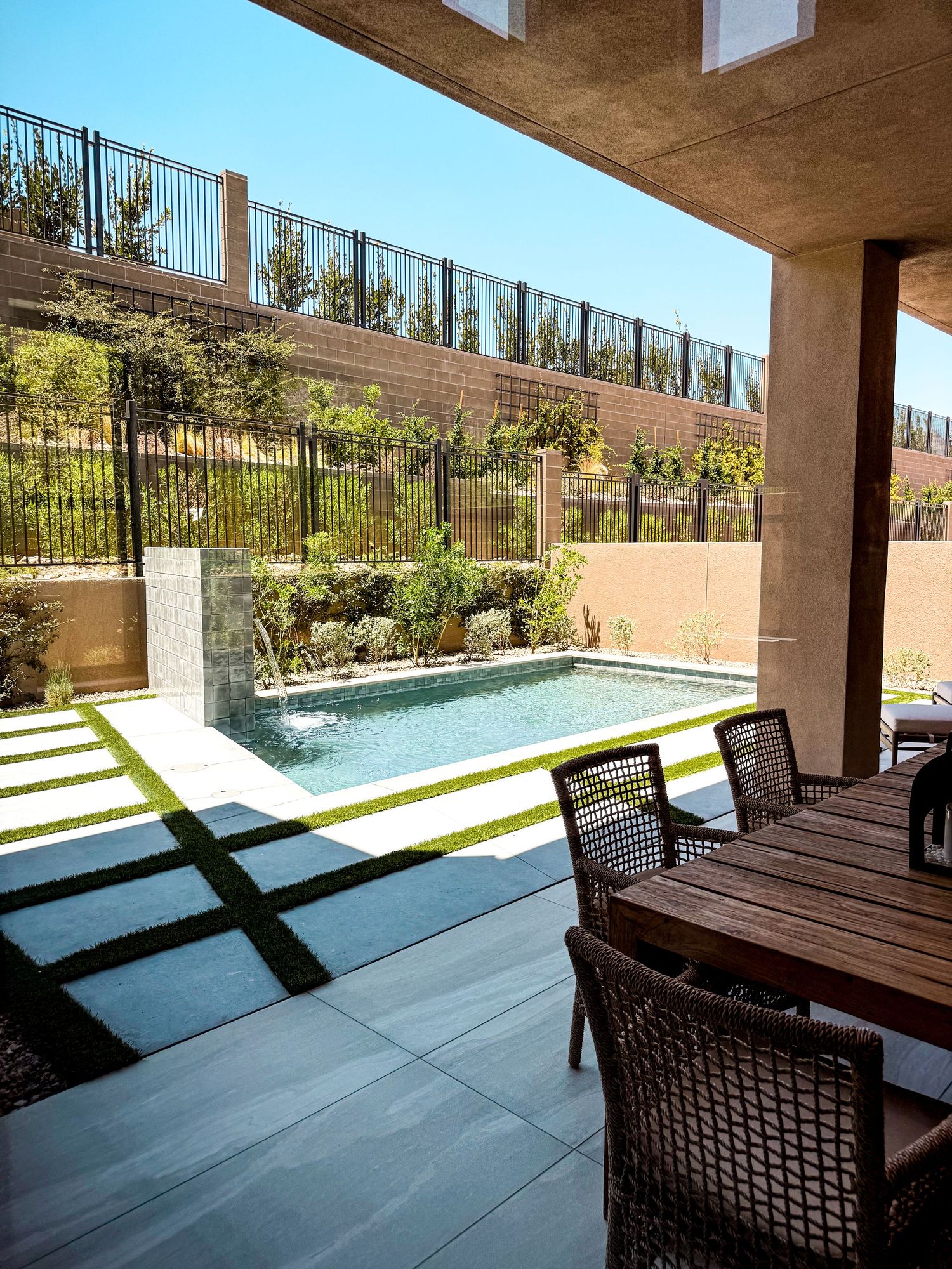 Modern backyard with rectangular pool, outdoor dining area, lush greenery, and iron fence under clear blue sky.