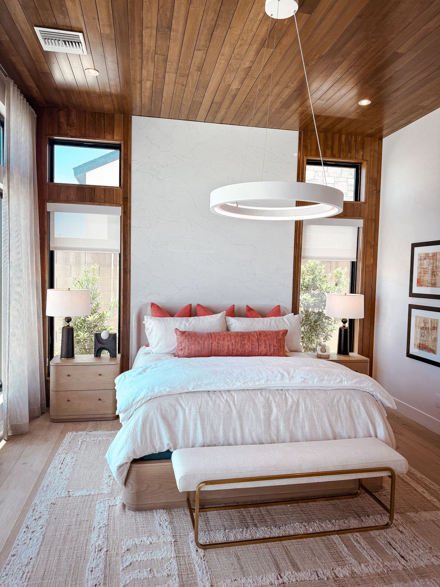 Modern bedroom with white bedding, coral pillows, wood ceiling, ring chandelier, and natural light from windows.