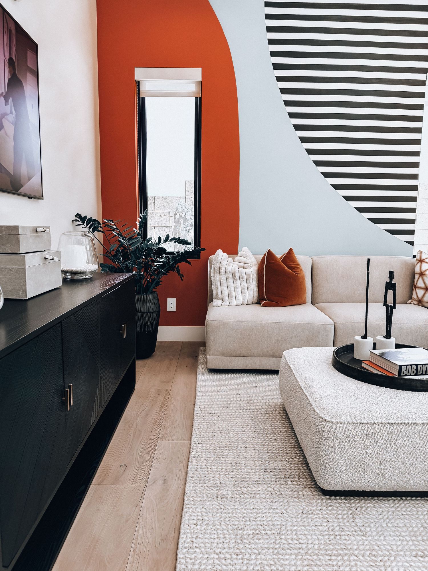 Modern living room with rust orange accent wall, striped mural, beige sectional sofa, black credenza, and neutral ottoman.