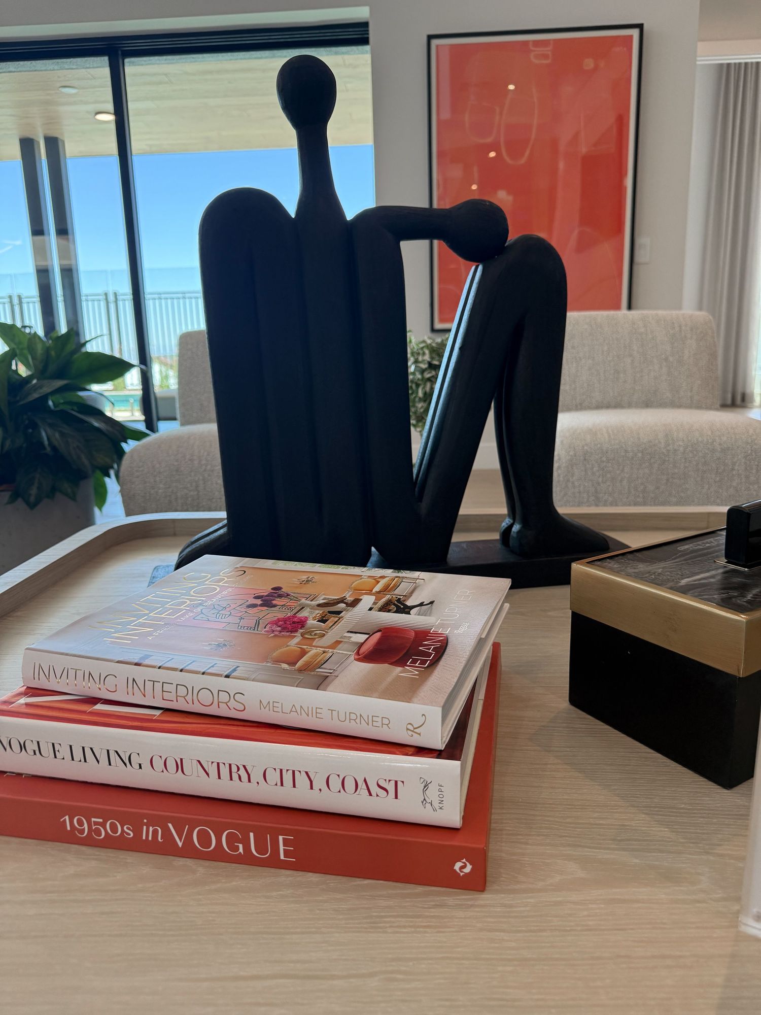 Black abstract sculpture on coffee table with Vogue and Interiors books, ocean view in modern living room.