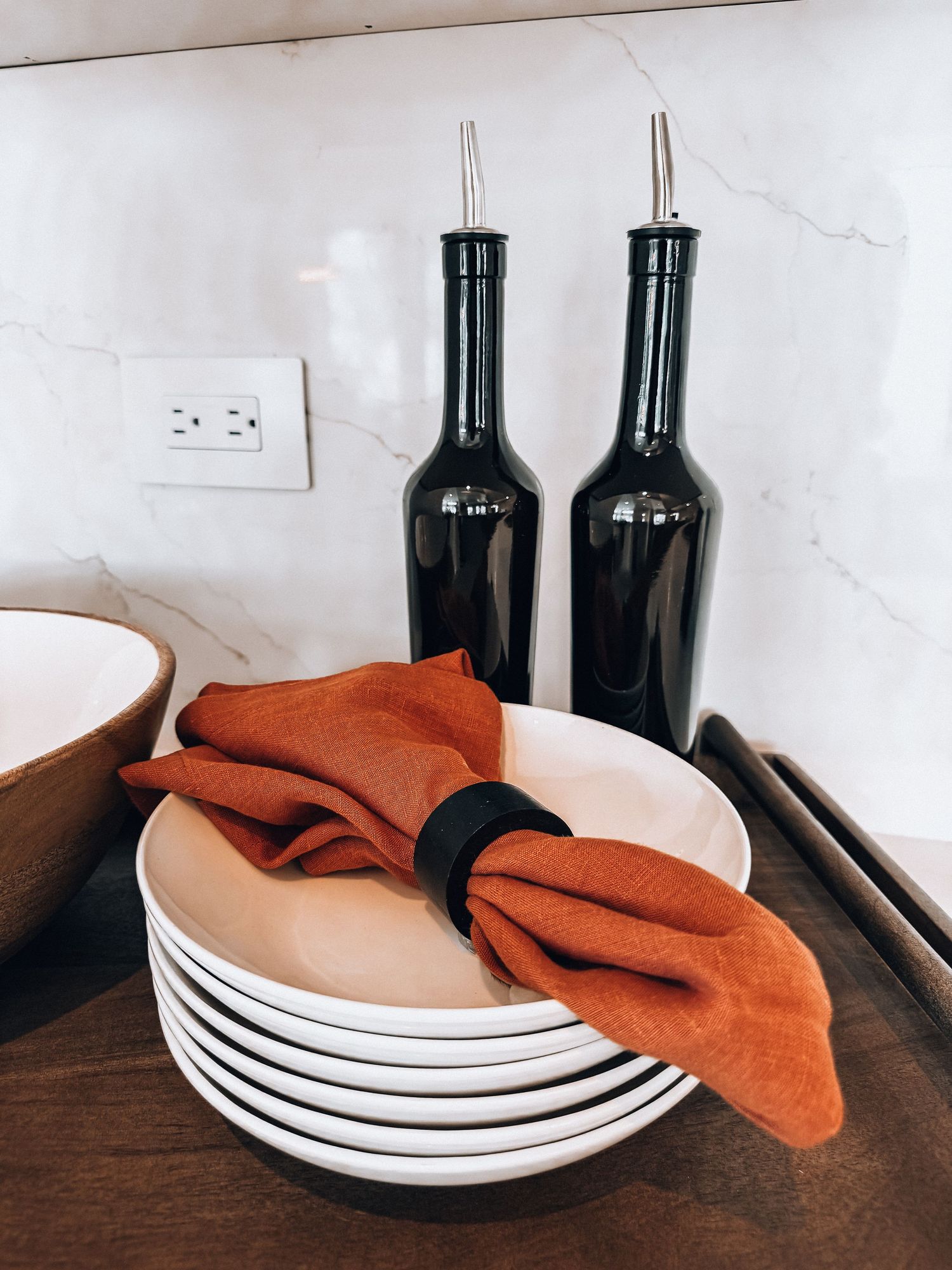 Stacked white plates with a rust orange linen napkin, wooden bowls, and dark glass oil bottles on a wooden tray.