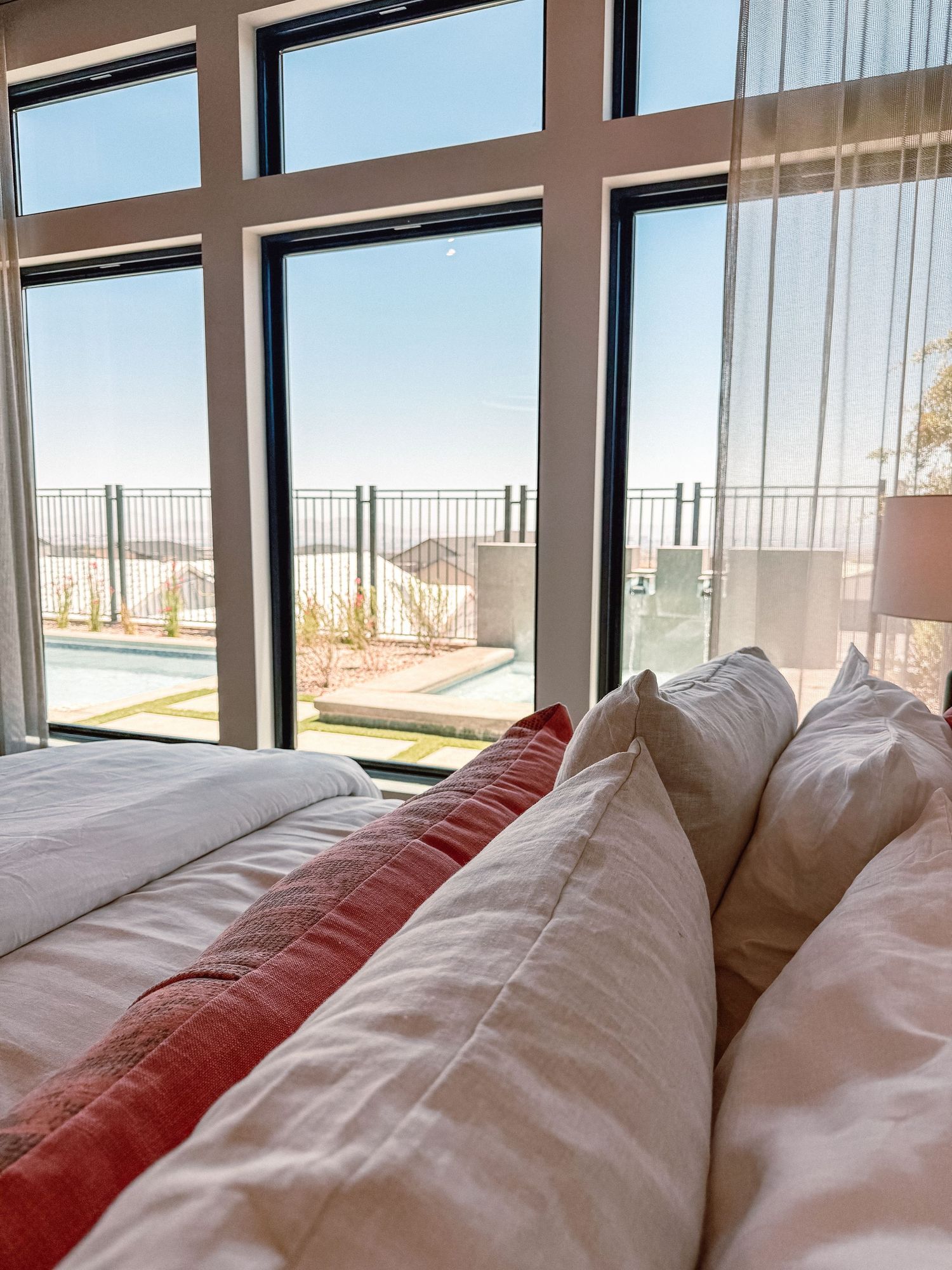 Bright bedroom with white and red pillows on a bed, large black-framed windows overlooking a pool and clear blue sky.