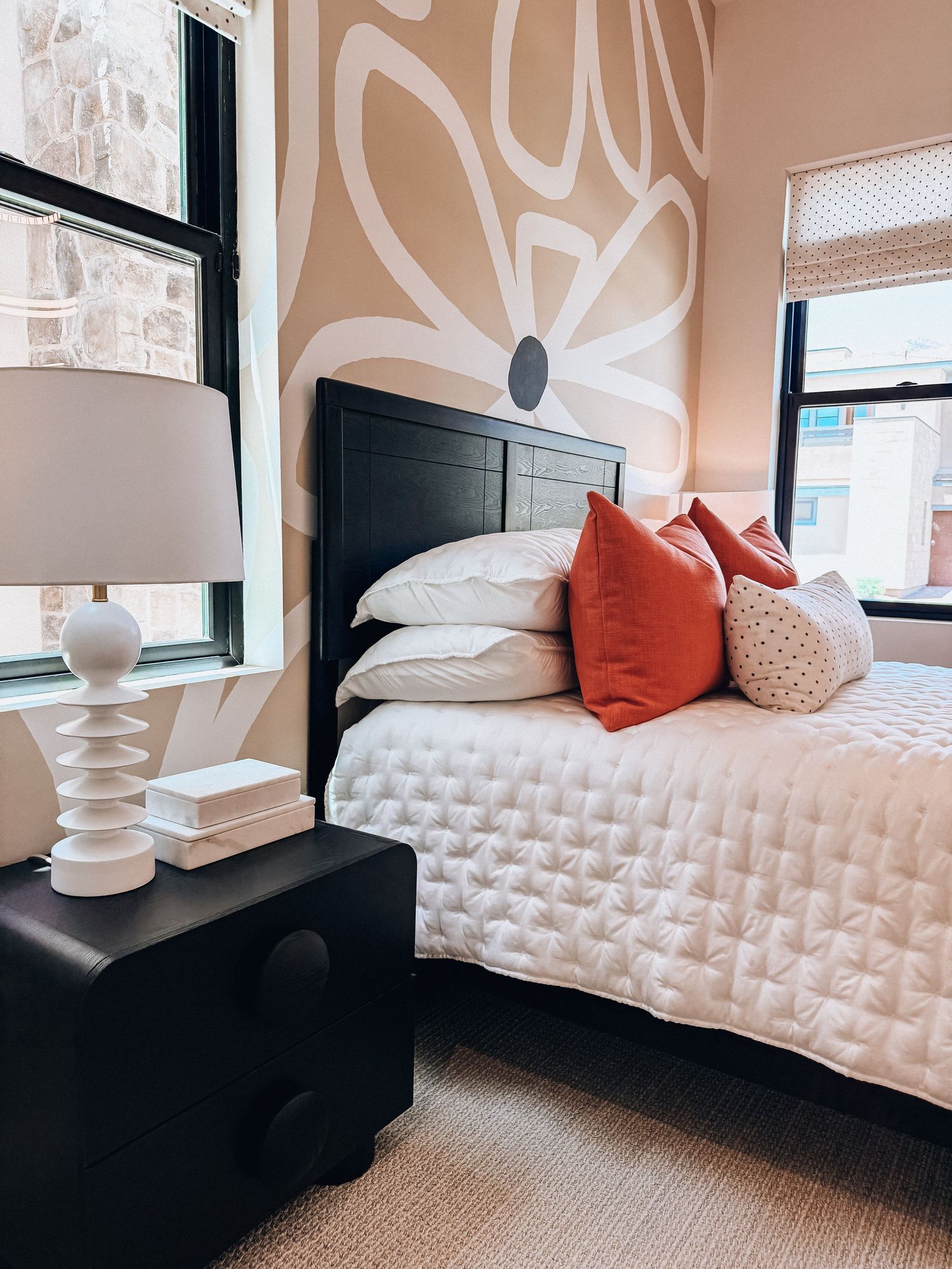 Modern bedroom with black headboard, white quilted bedding, coral pillows, abstract floral mural wall, and white geometric lamp.