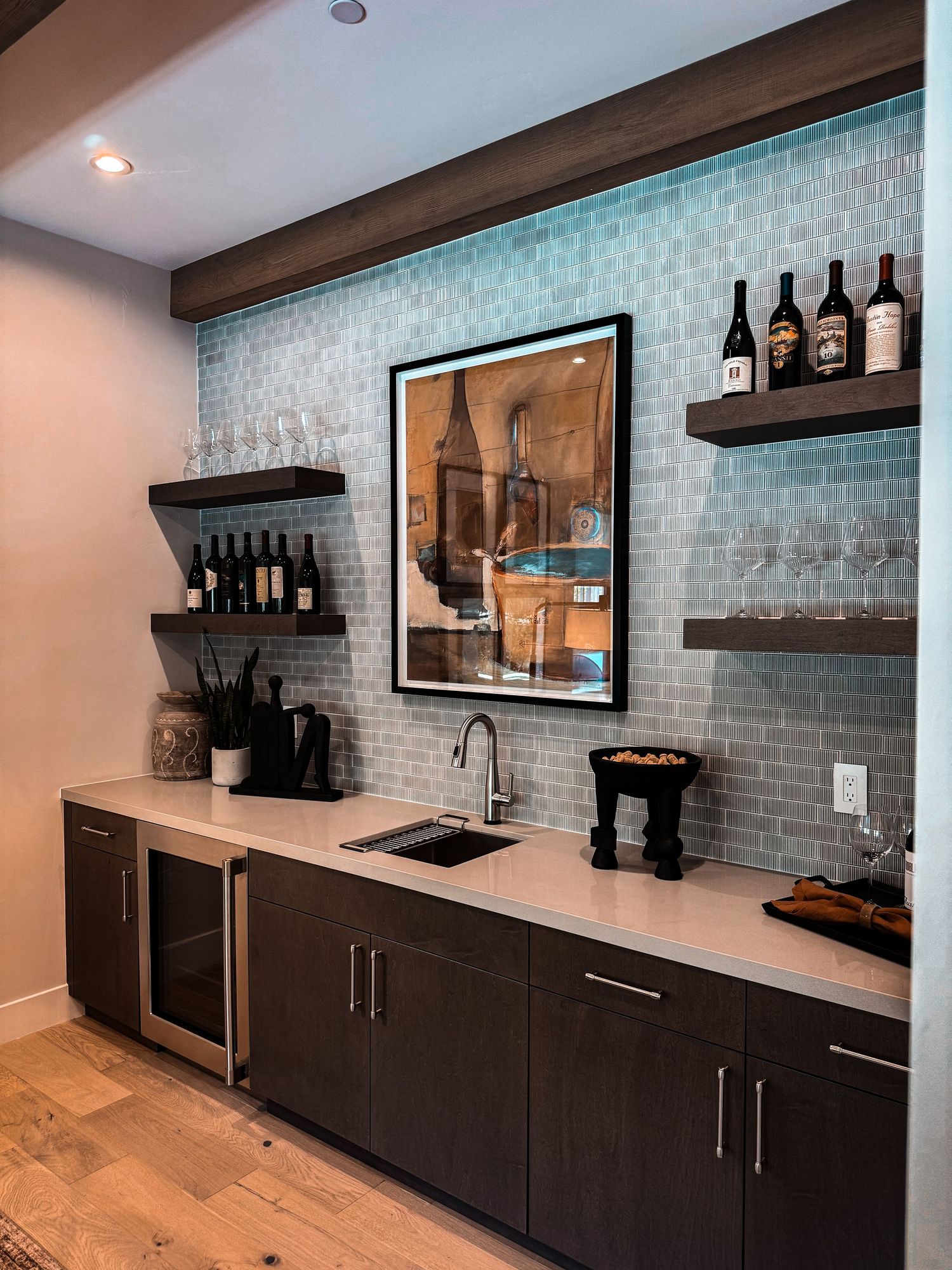 Modern home bar with teal mosaic tile backsplash, dark floating shelves with wine bottles, and white quartz countertop.