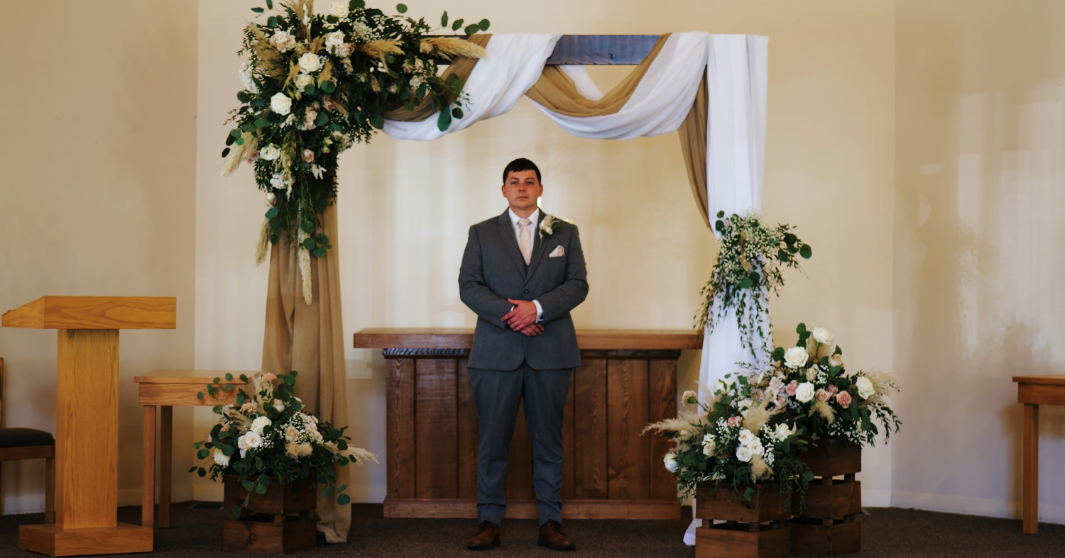 Groom in a gray suit standing at the floral arch altar before the wedding ceremony begins.