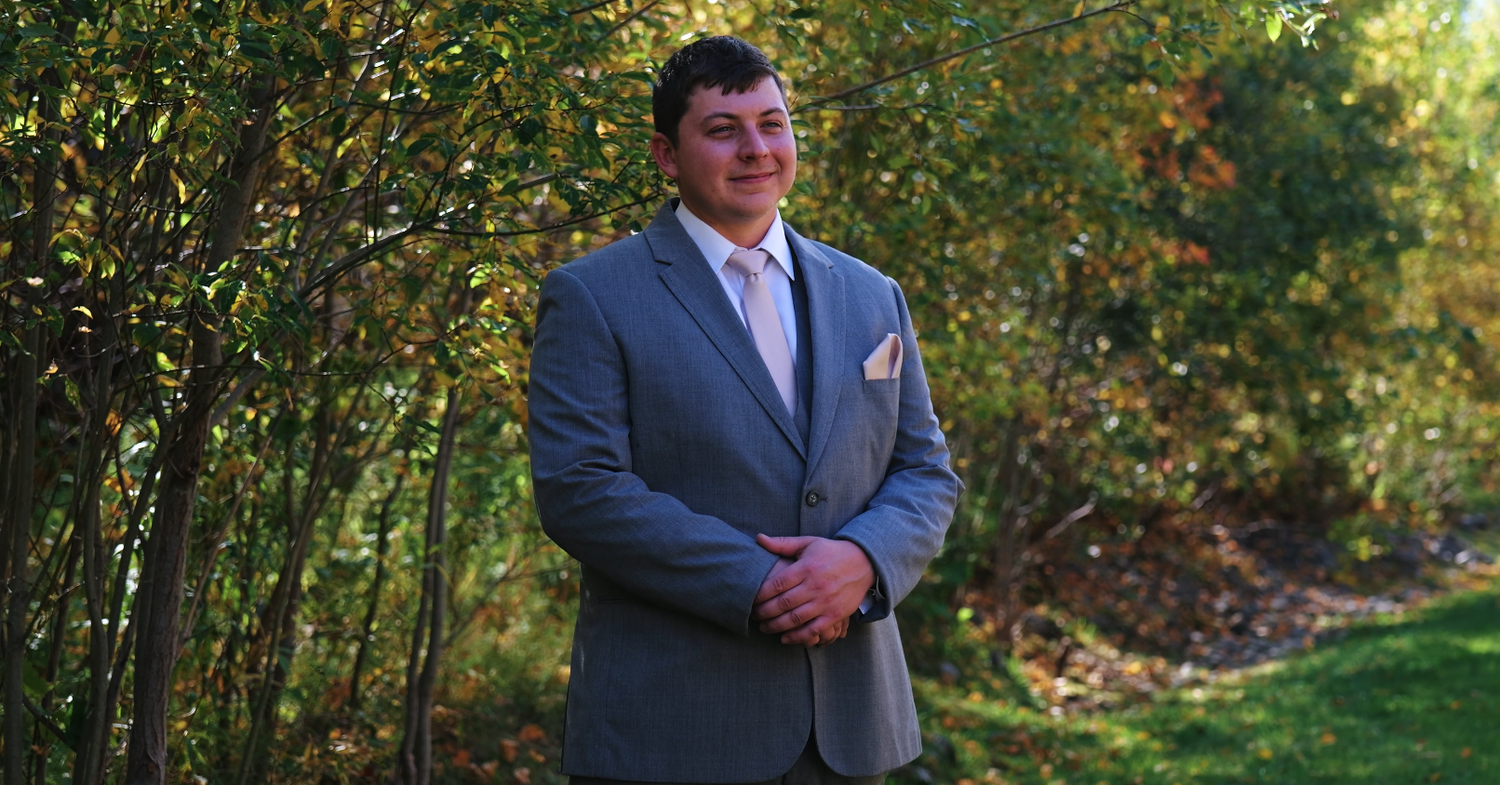 Groomsman in a gray suit posing outdoors surrounded by lush autumn foliage.