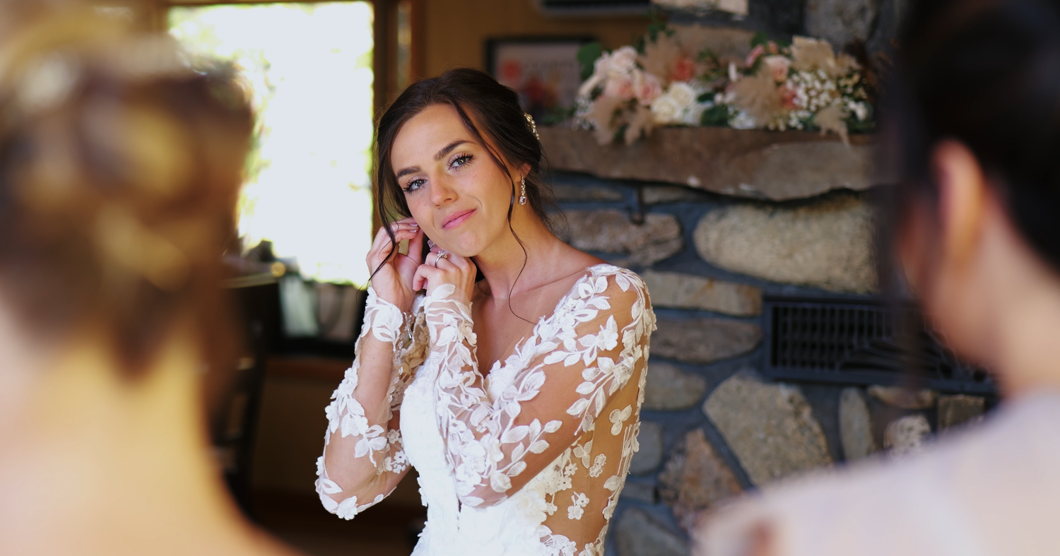 Bride in a lace wedding gown touching her face and smiling in front of a stone fireplace.