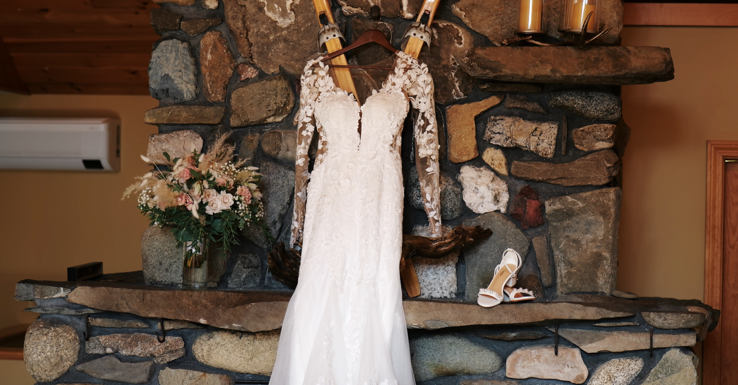 Lace wedding dress hanging on a wooden hanger in front of a stone fireplace.