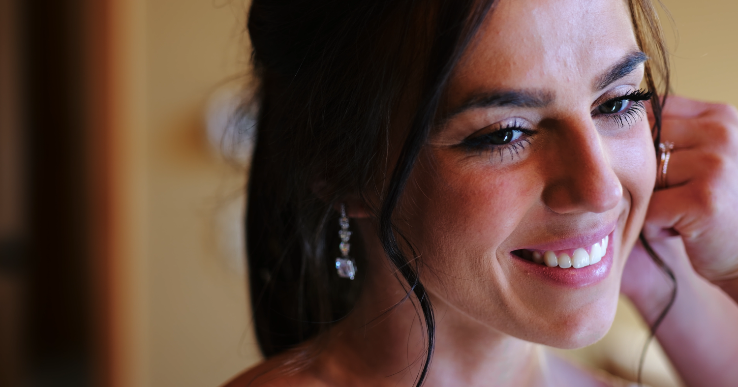 Bride smiling while putting on a crystal drop earring during wedding day preparations.