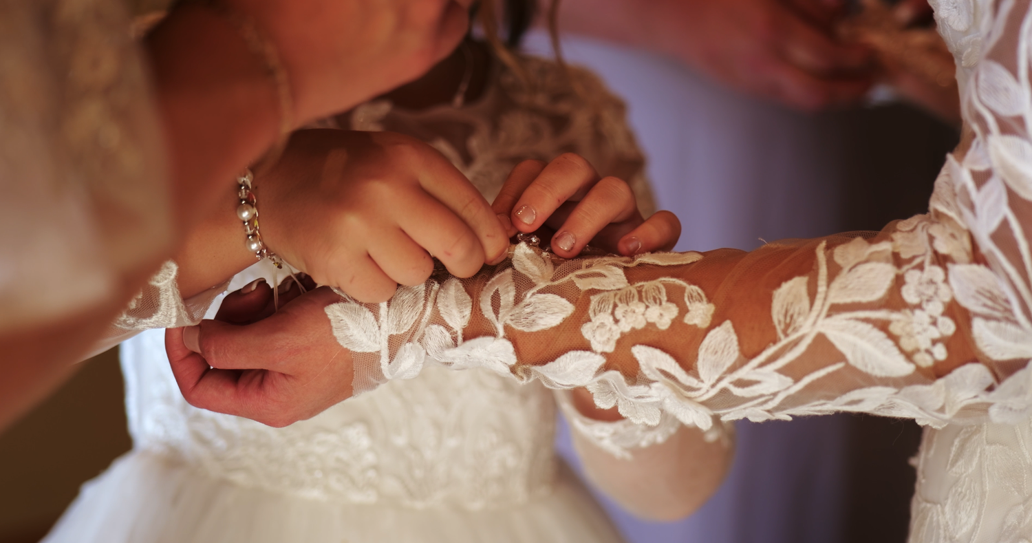 Bridesmaid fastening the lace buttons on the back of the bride's long-sleeve wedding gown.