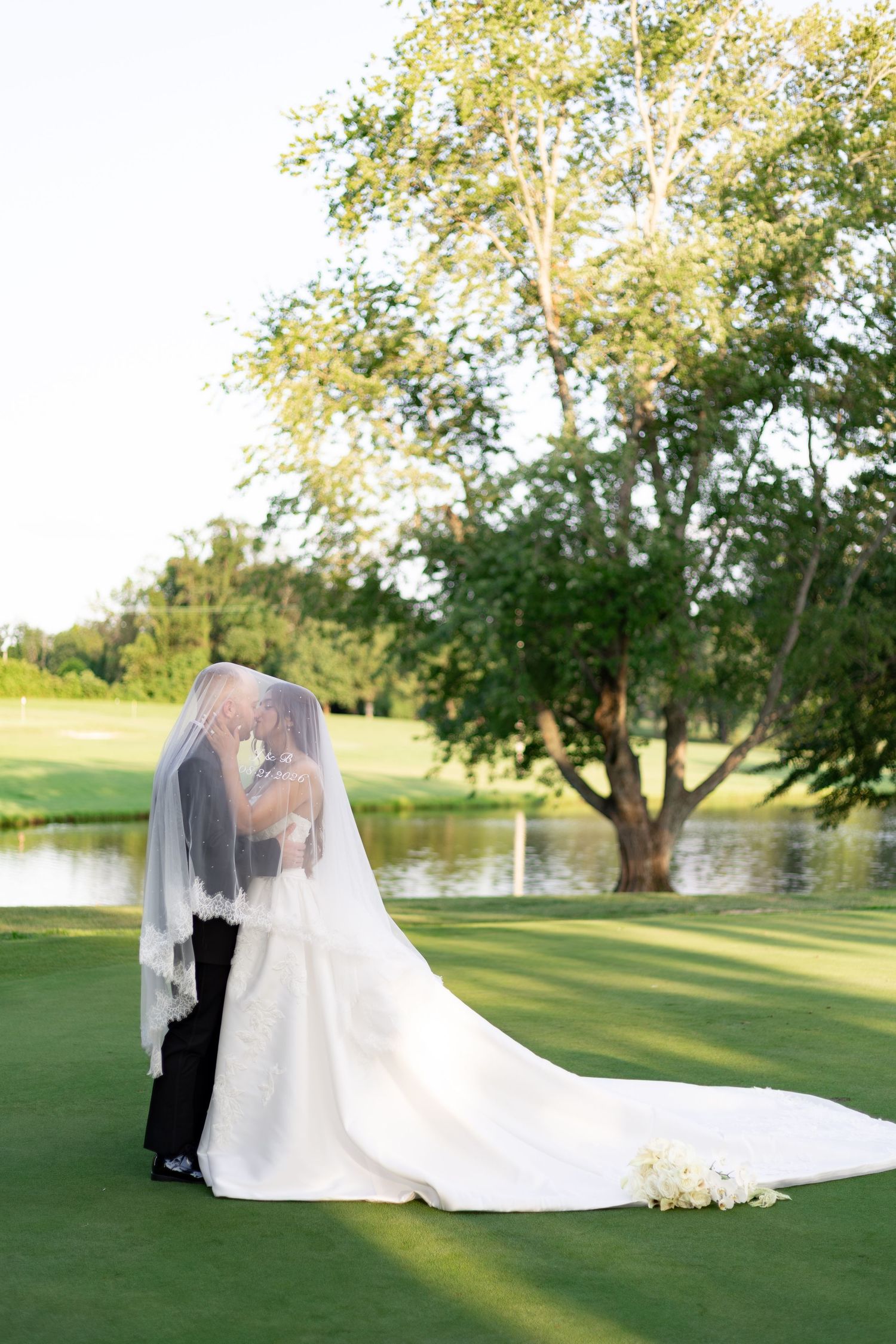 Bride and groom embrace under a long white veil on a sunlit golf course lawn with a tree and calm pond behind them.