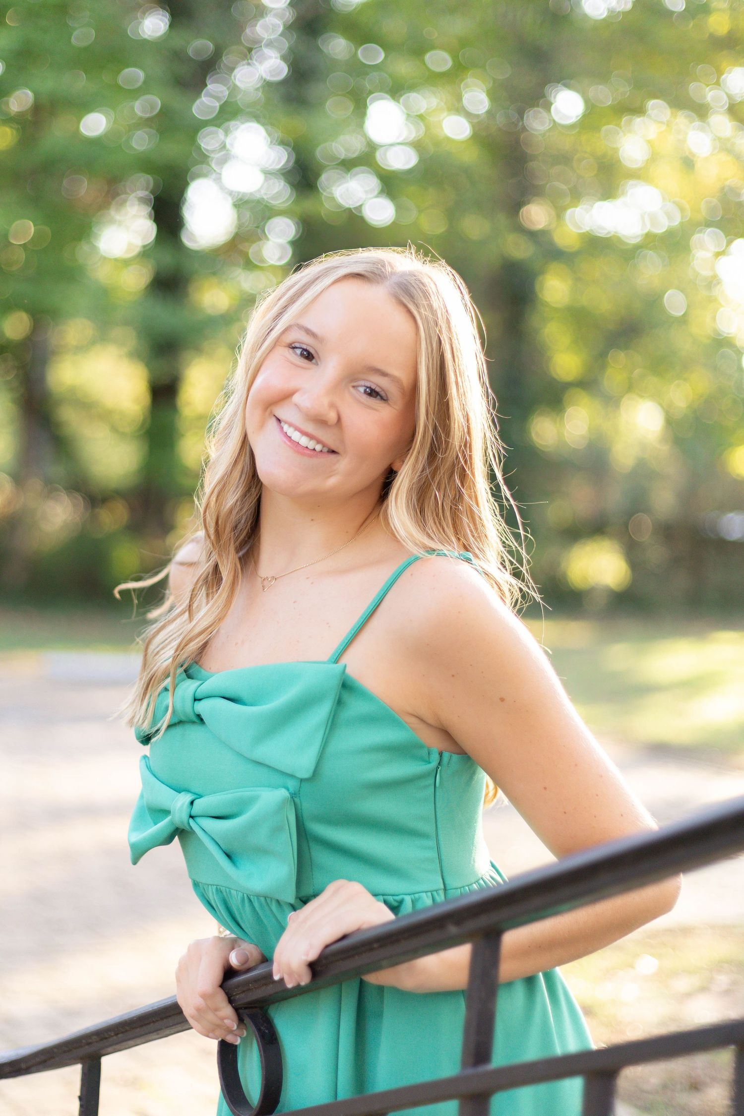 Happy blonde teen girl in teal bow dress smiling while leaning on black railing in sunny park.