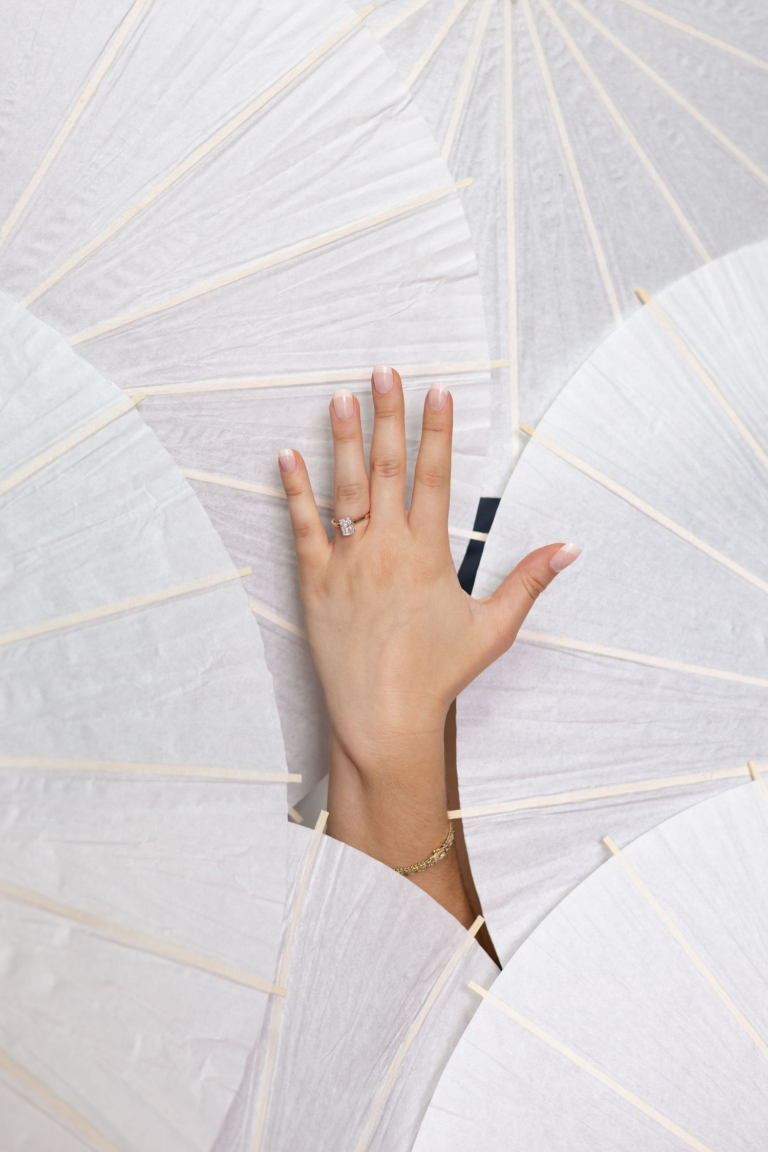 A delicate hand with a diamond ring and gold bracelet rests gracefully among white paper parasols.