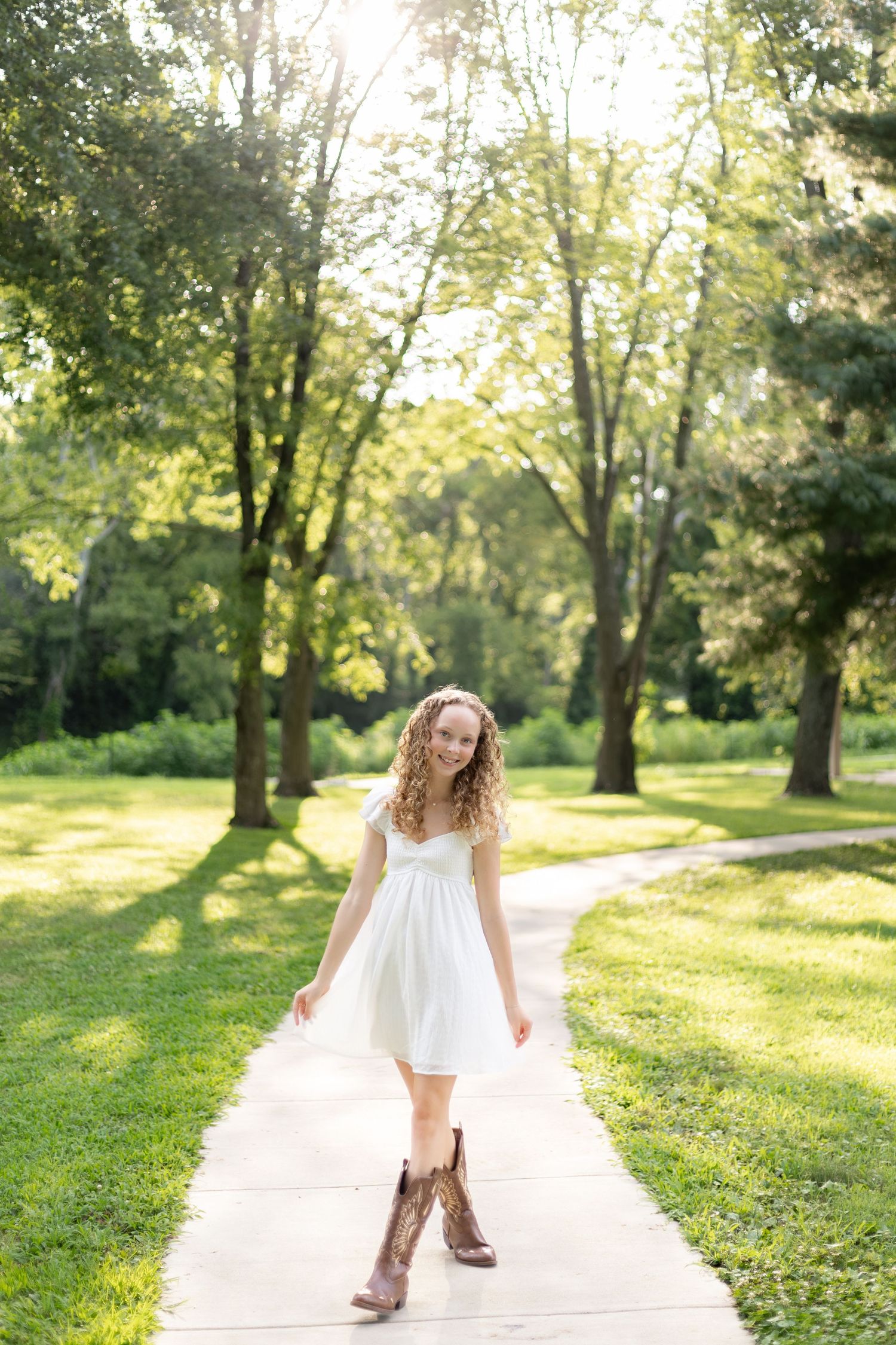 Young woman in white dress and cowboy boots smiling on a sunlit park path surrounded by lush green trees.
