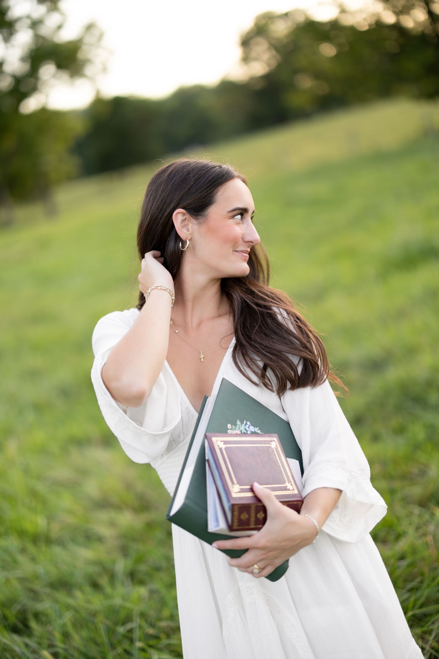 A smiling brunette woman in a white dress holds green books in a sunlit grassy field.