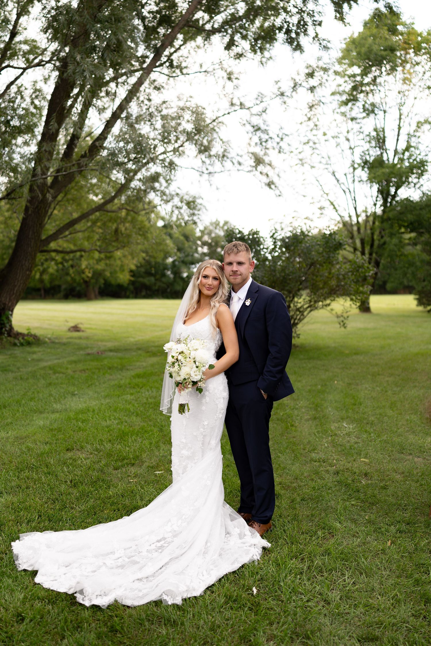 Bride in lace gown with bouquet and groom in navy suit pose together on green lawn surrounded by trees.
