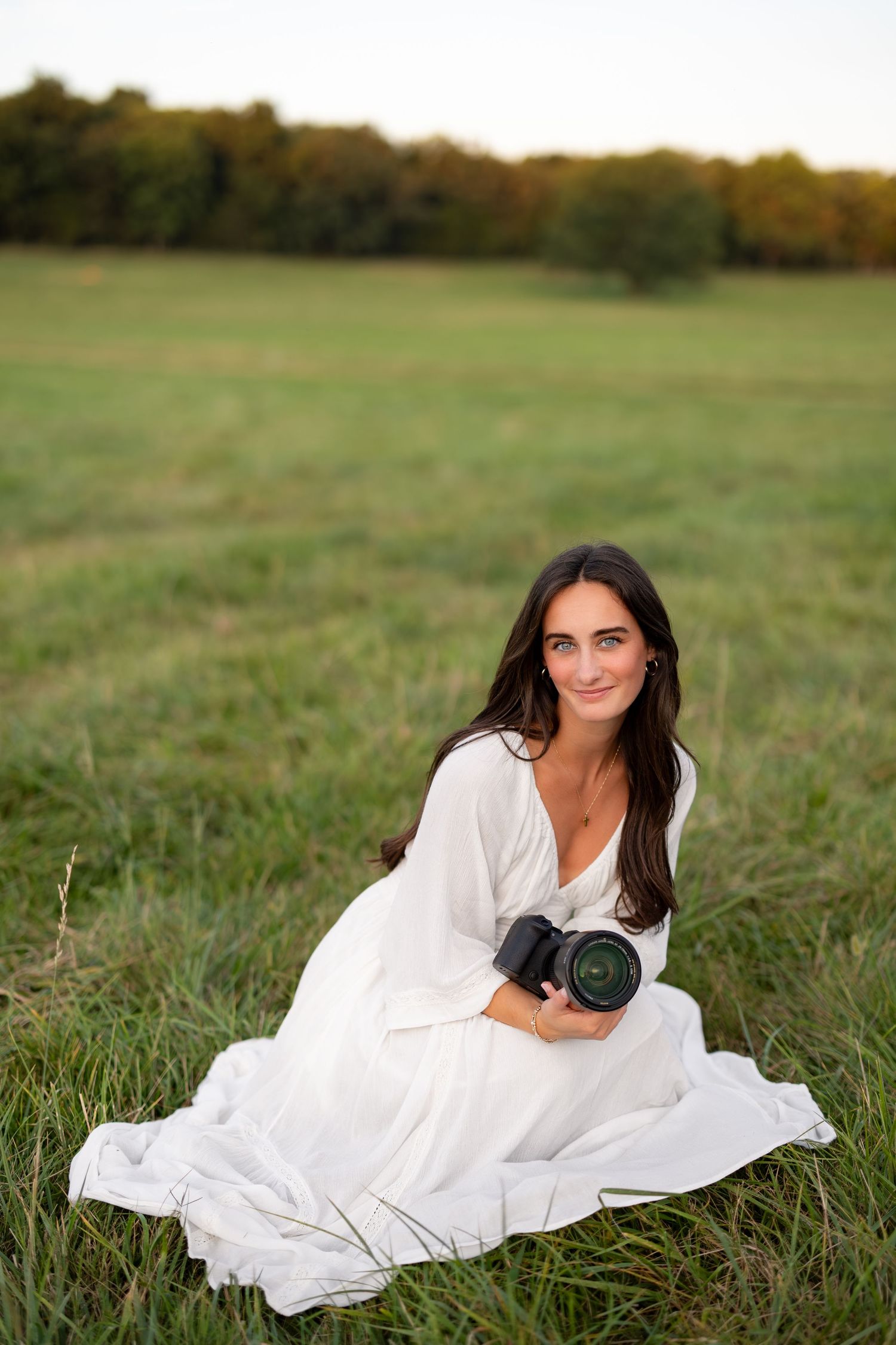 Woman in white dress sitting in green field holding DSLR camera, smiling at camera with trees in background.