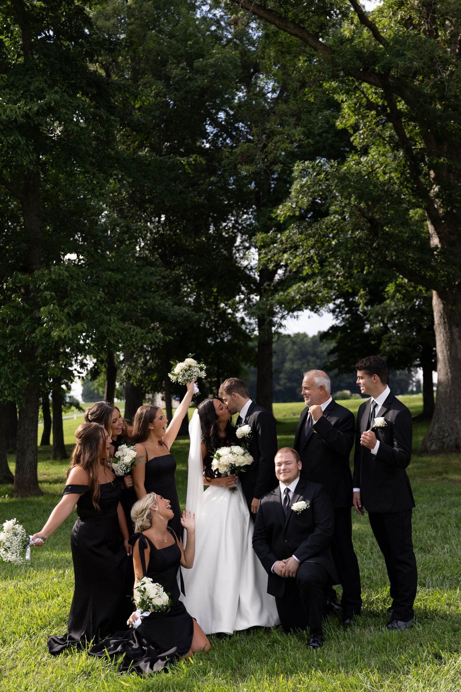 Wedding party in black attire surrounds bride and groom outdoors, with bridesmaids holding white bouquets among lush green trees.
