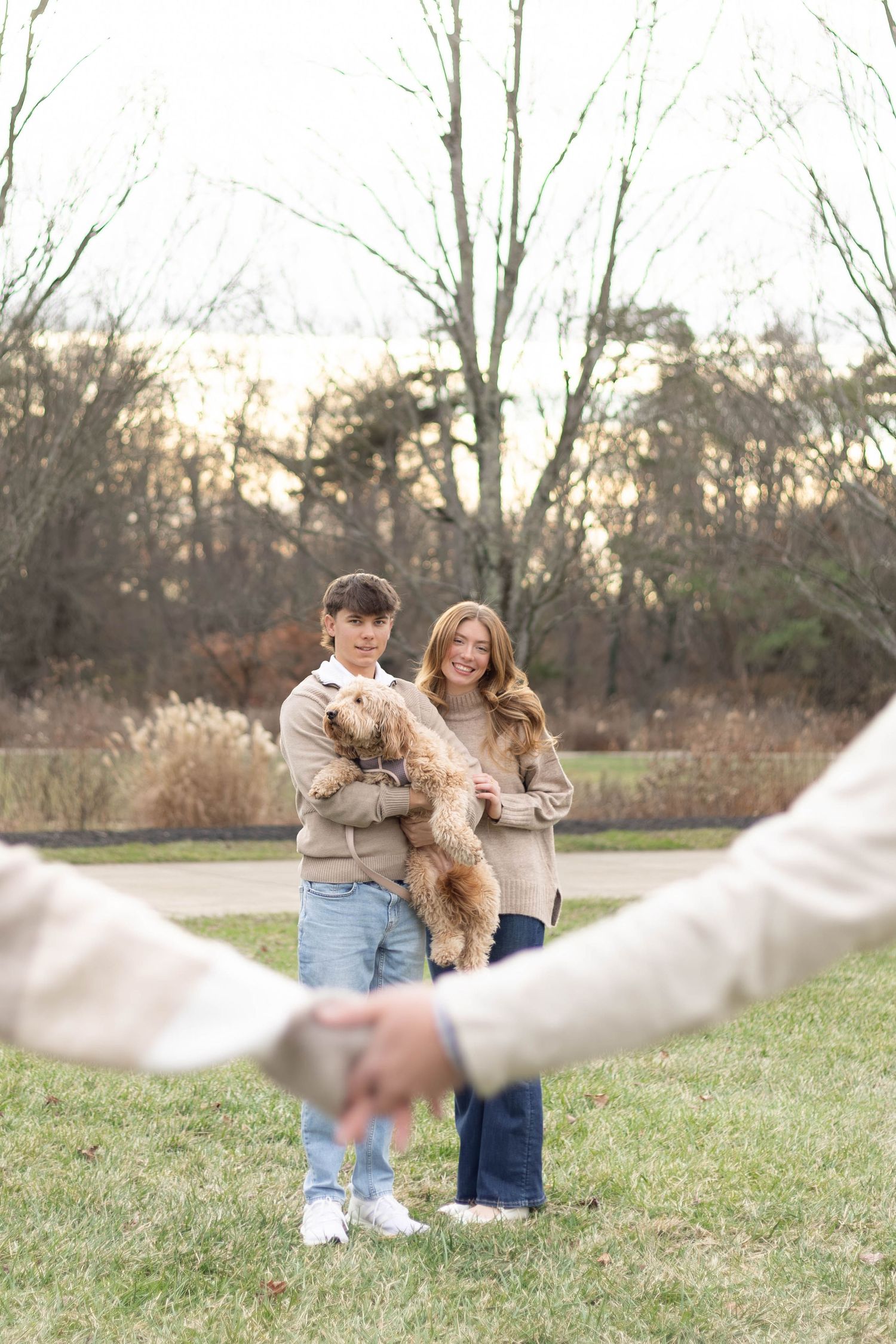 A young couple holds a fluffy dog in a park at dusk, framed by linked hands.