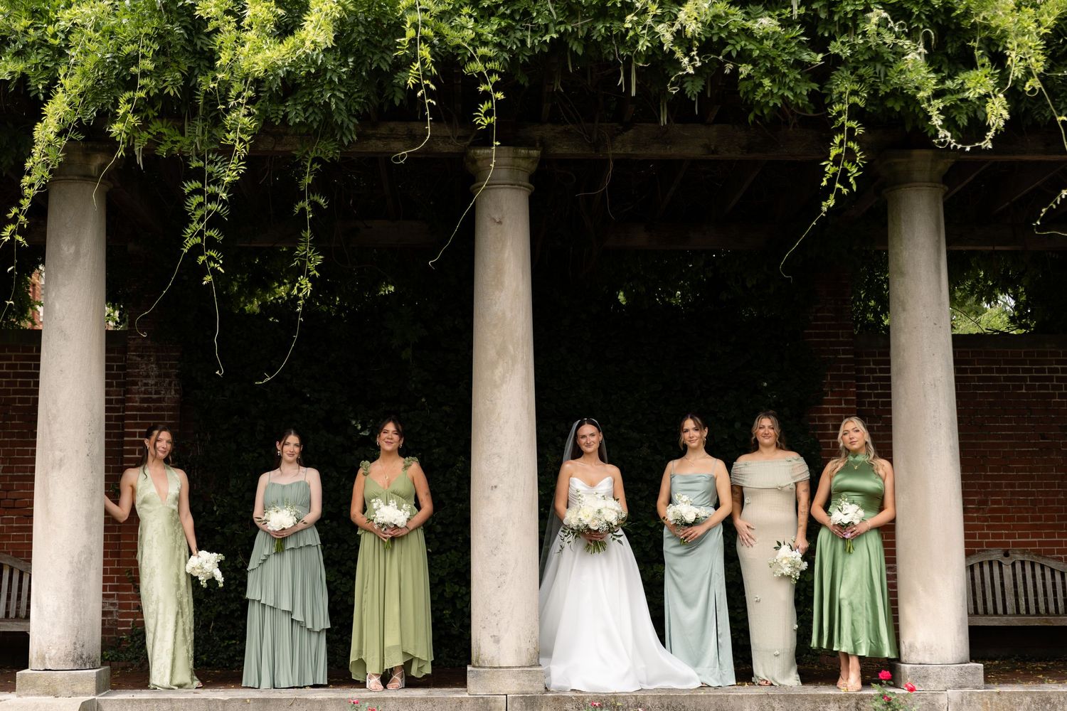 Bridesmaids in mixed sage green gowns stand with bride under vine-draped stone columns.