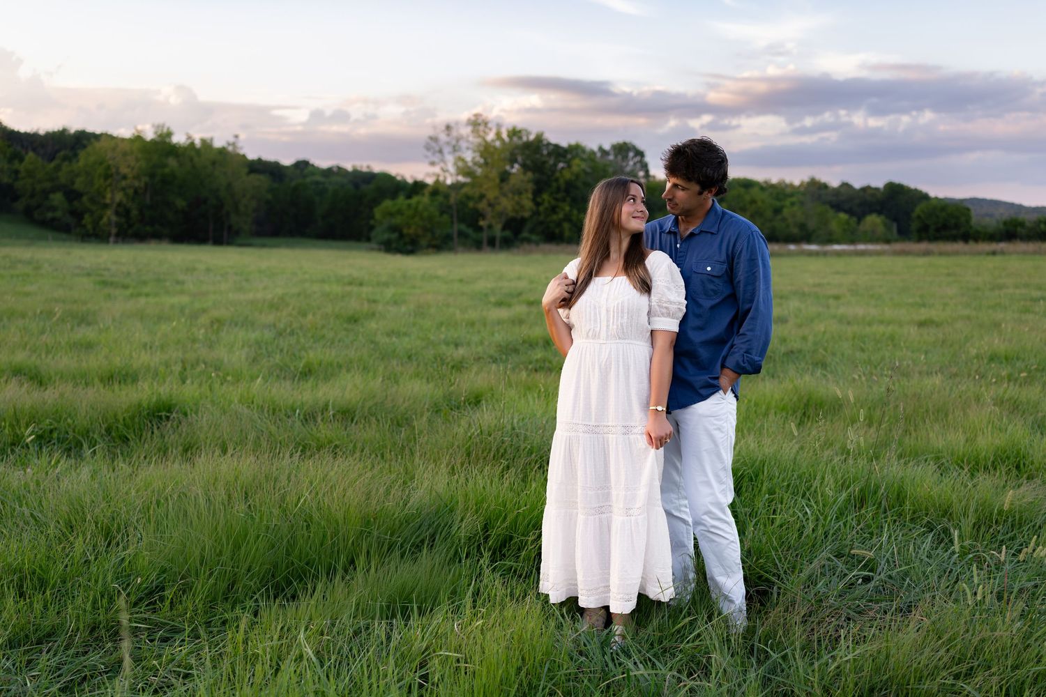 Couple standing in a lush green field at sunset, woman in white dress and man in blue shirt.
