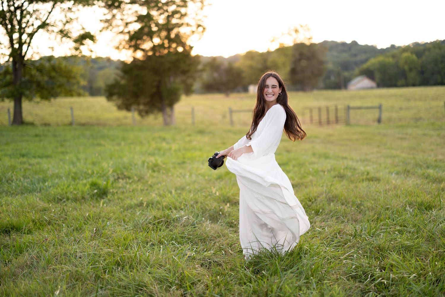 Young woman in white dress holding camera twirls in sunlit green field at golden hour near wooden fence.