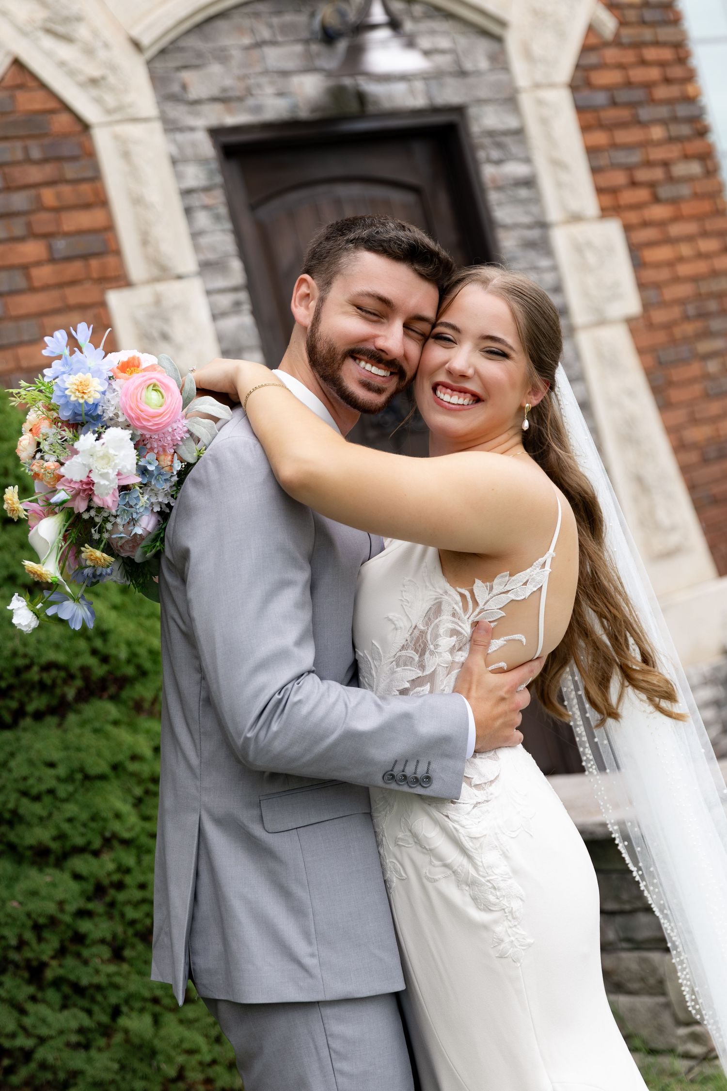 Smiling bride and groom embrace outside a stone and brick church, she holds a pastel floral bouquet and wears a lace gown with veil.