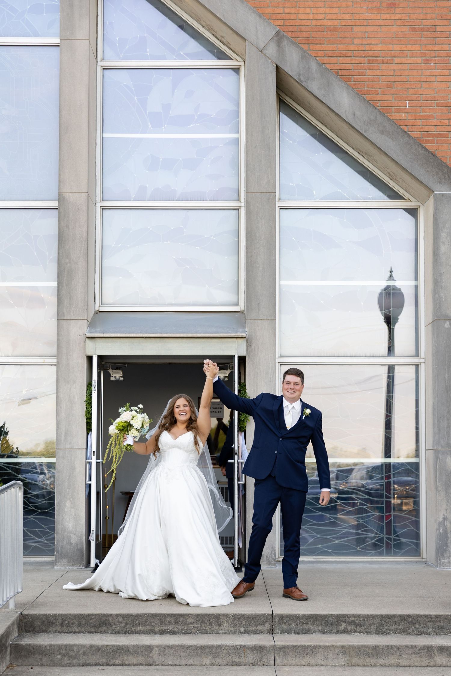 Bride and groom celebrating on church steps, raising hands in joy, bride in white gown holding bouquet, groom in navy suit.