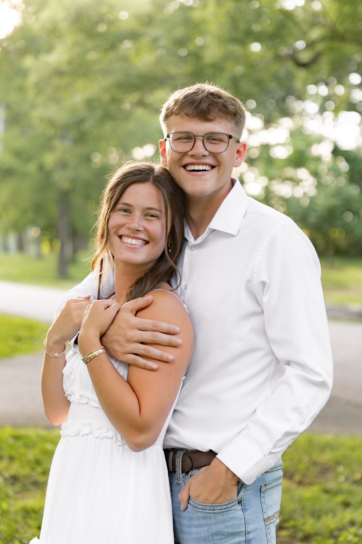 Happy young couple embracing outdoors, man in white shirt with glasses hugging smiling woman in white dress.