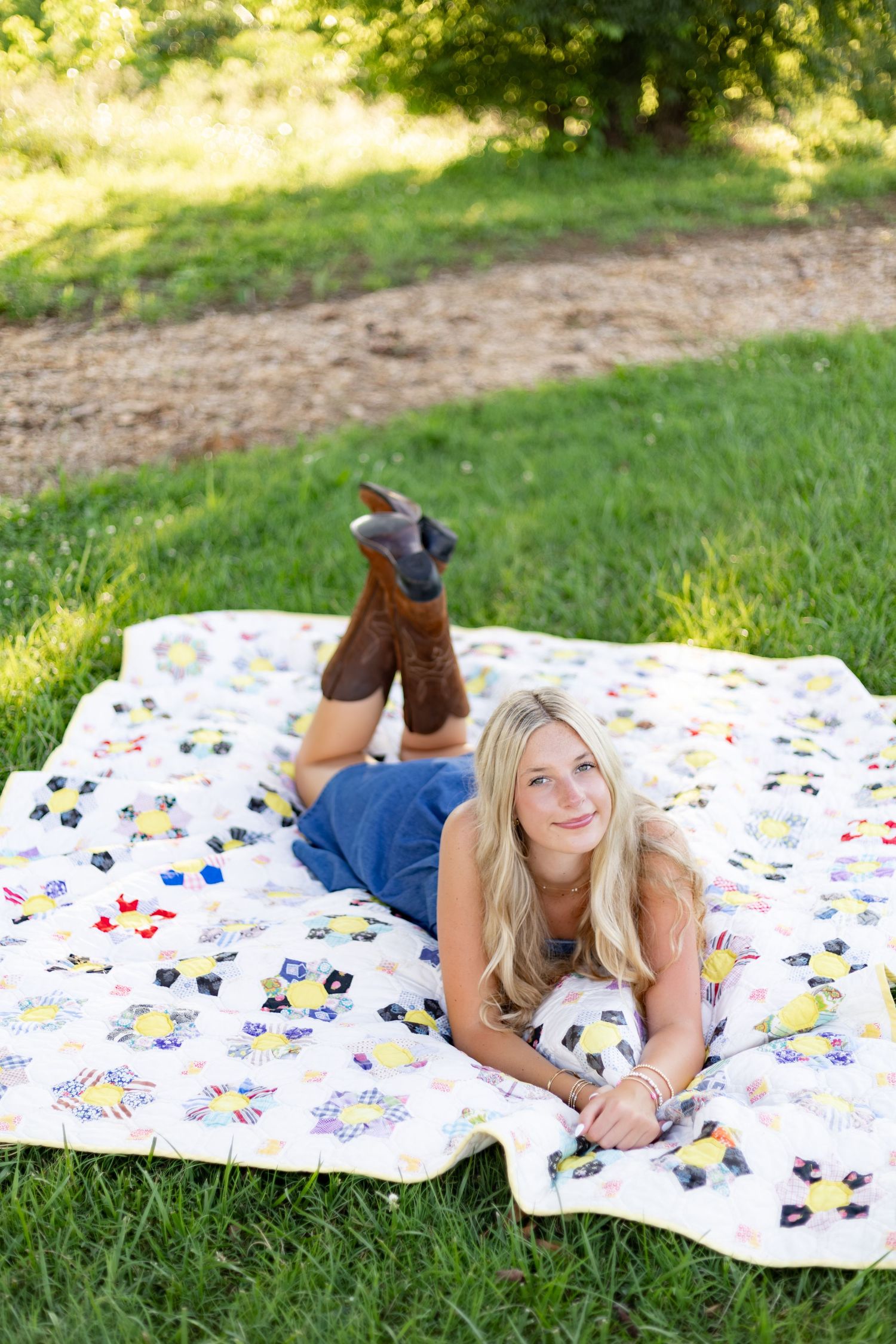 Young blonde woman lying on a patchwork quilt in green grass, wearing jeans and cowboy boots, smiling.