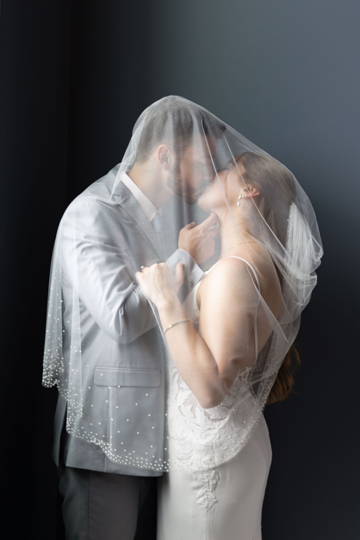 A bride and groom share a kiss beneath a sheer beaded wedding veil against a dark backdrop, the groom in a light gray suit.