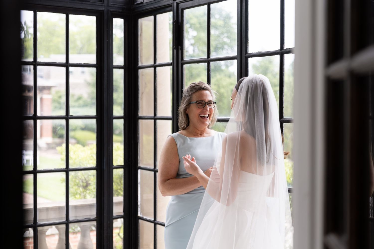 A bride and her mother share a joyful moment by elegant French doors before the wedding.