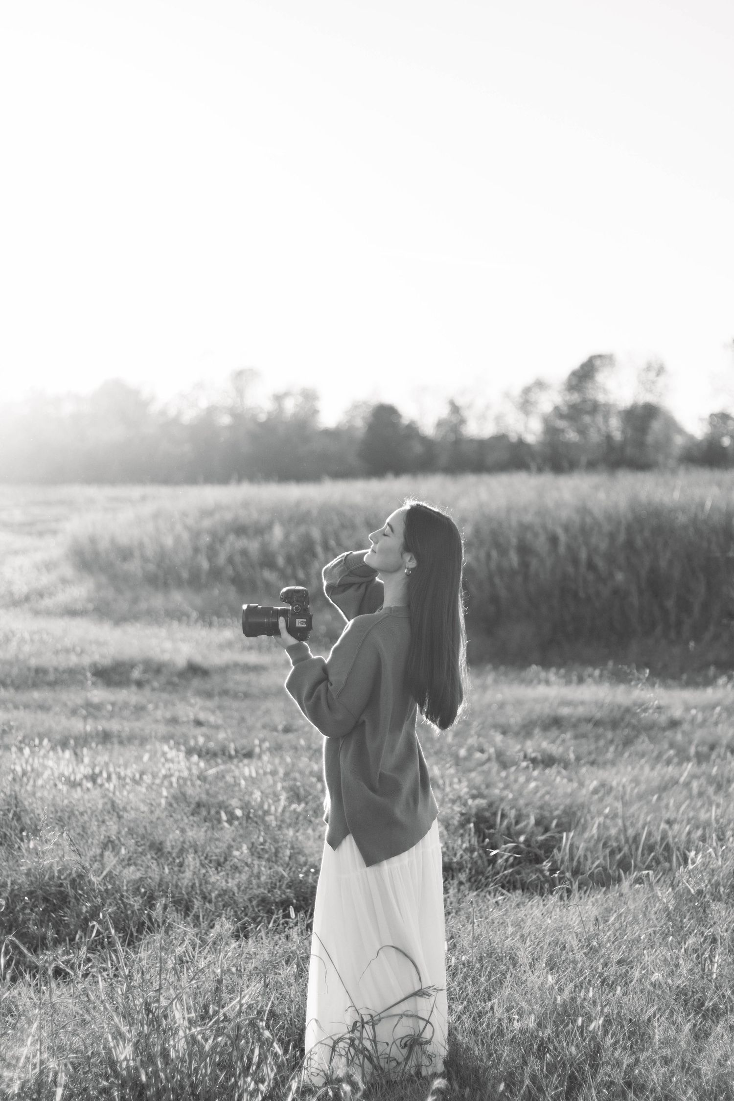 Black and white photo of a woman holding a camera in a field at sunset.
