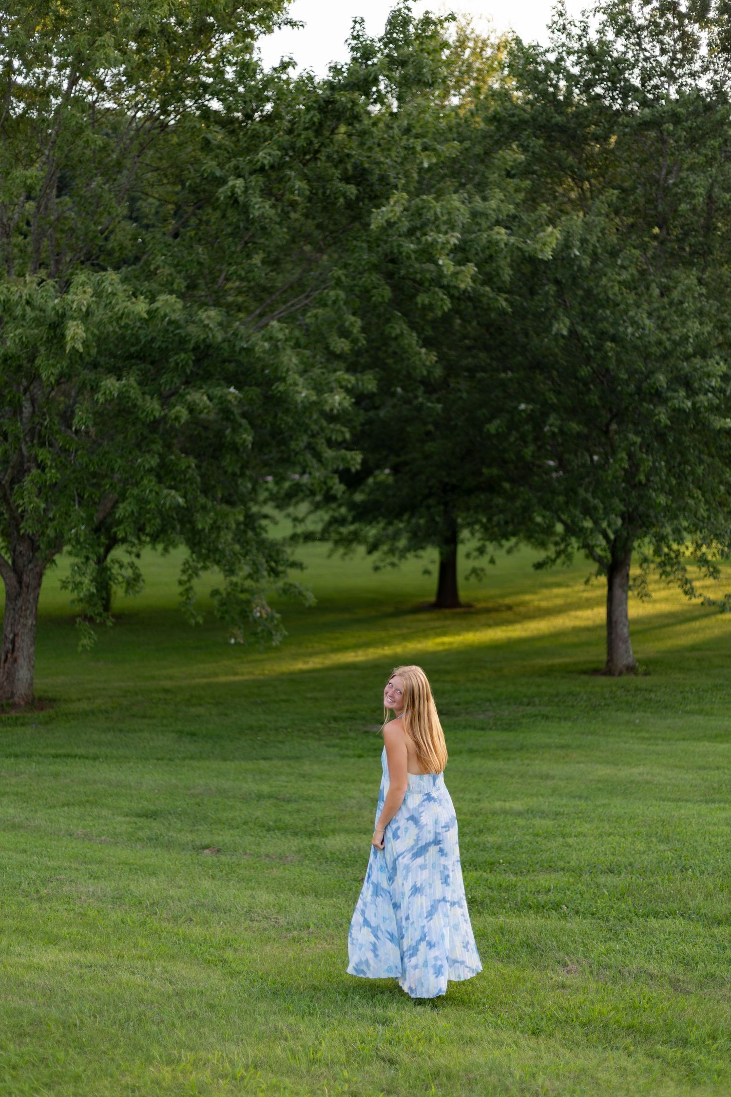 Young woman in blue floral maxi dress standing on lush green grass in a park surrounded by trees.