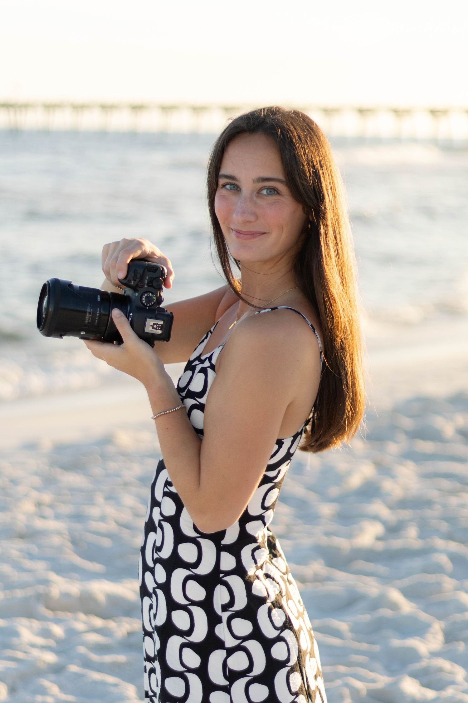 Young woman in black and white dress holding DSLR camera on sandy beach at sunset.