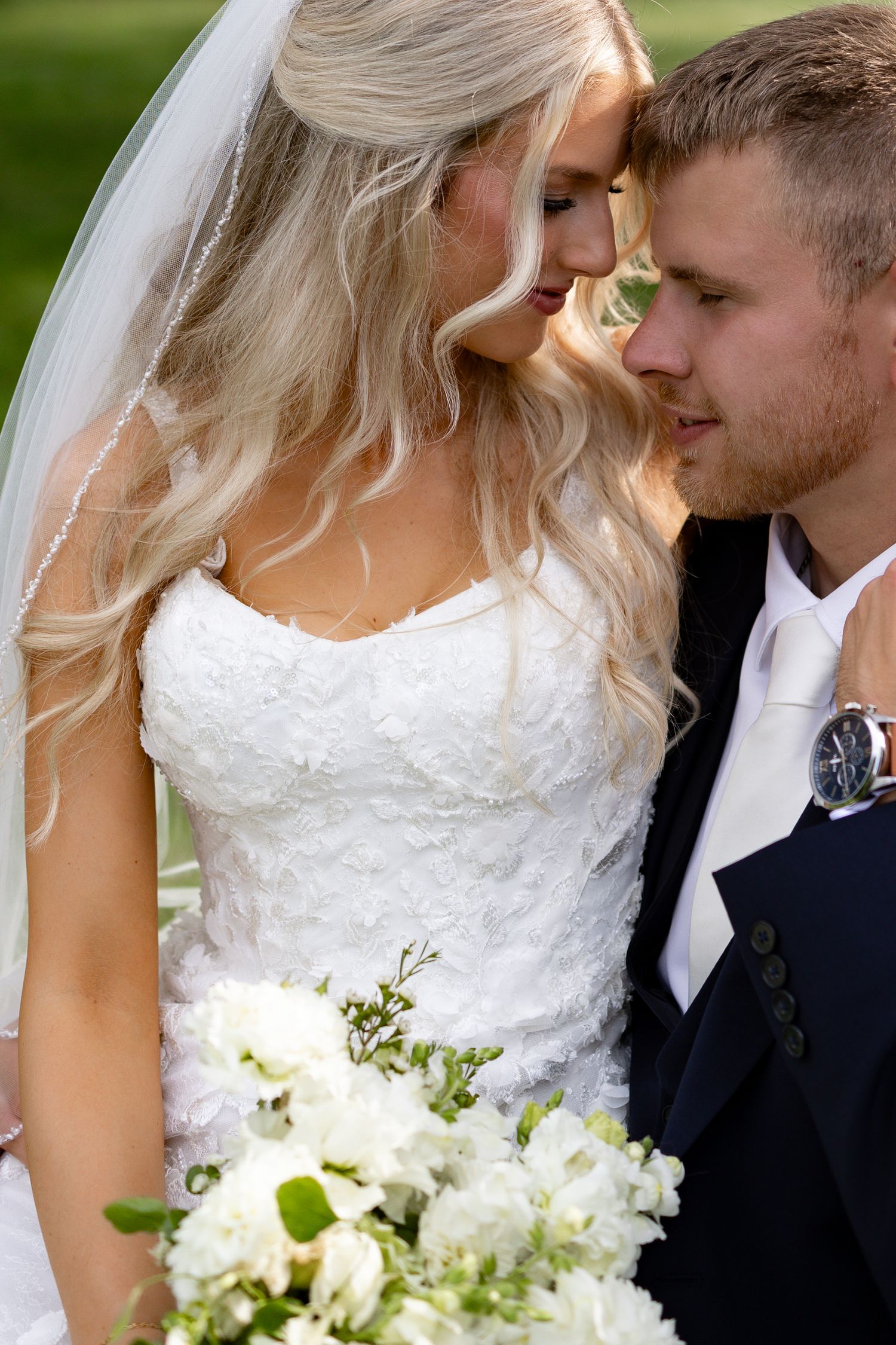Bride and groom touching foreheads tenderly, bride in white lace gown holding white floral bouquet outdoors.