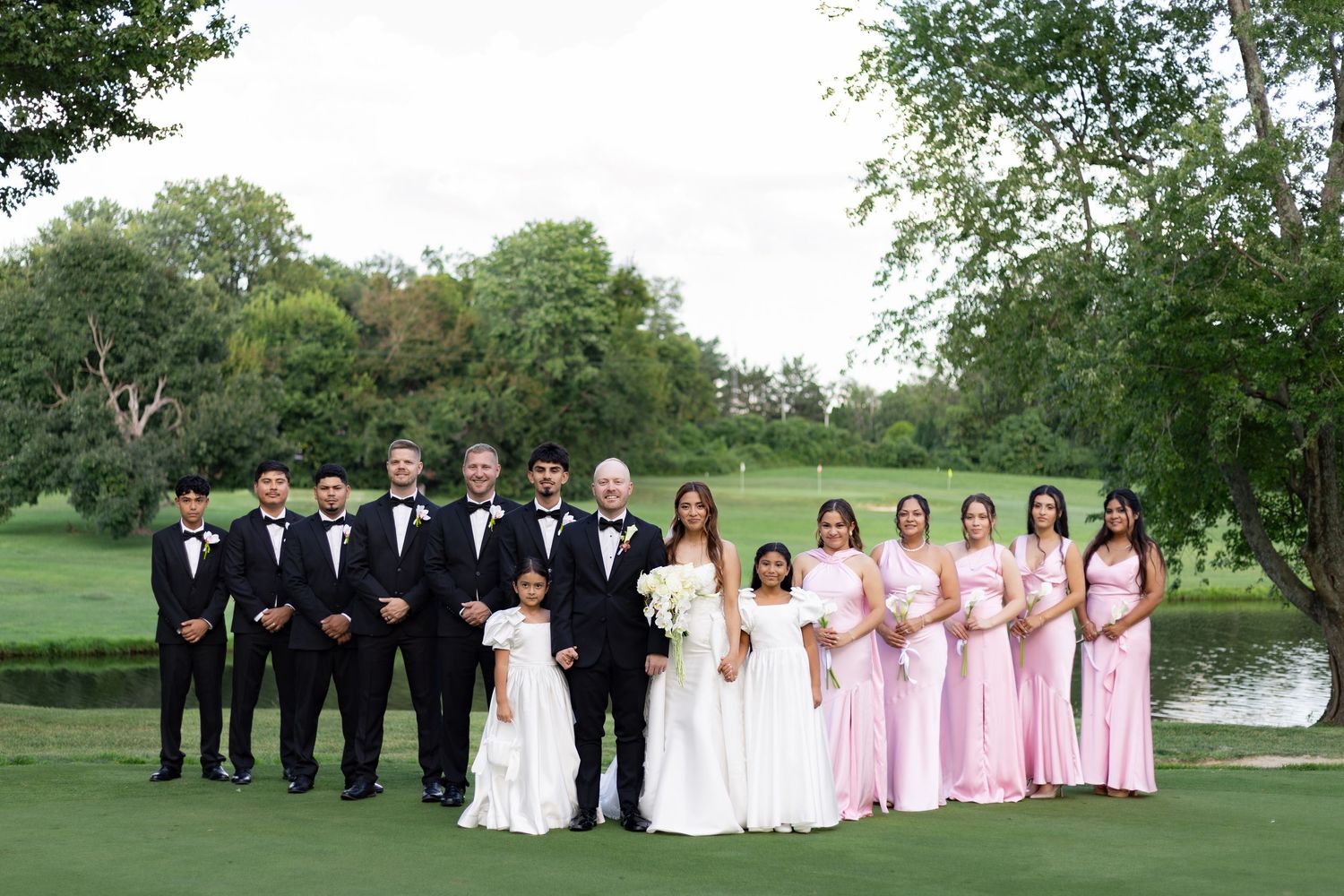 Wedding party poses outdoors on golf course, groomsmen in tuxedos, bridesmaids in pink gowns, bride and groom at center.