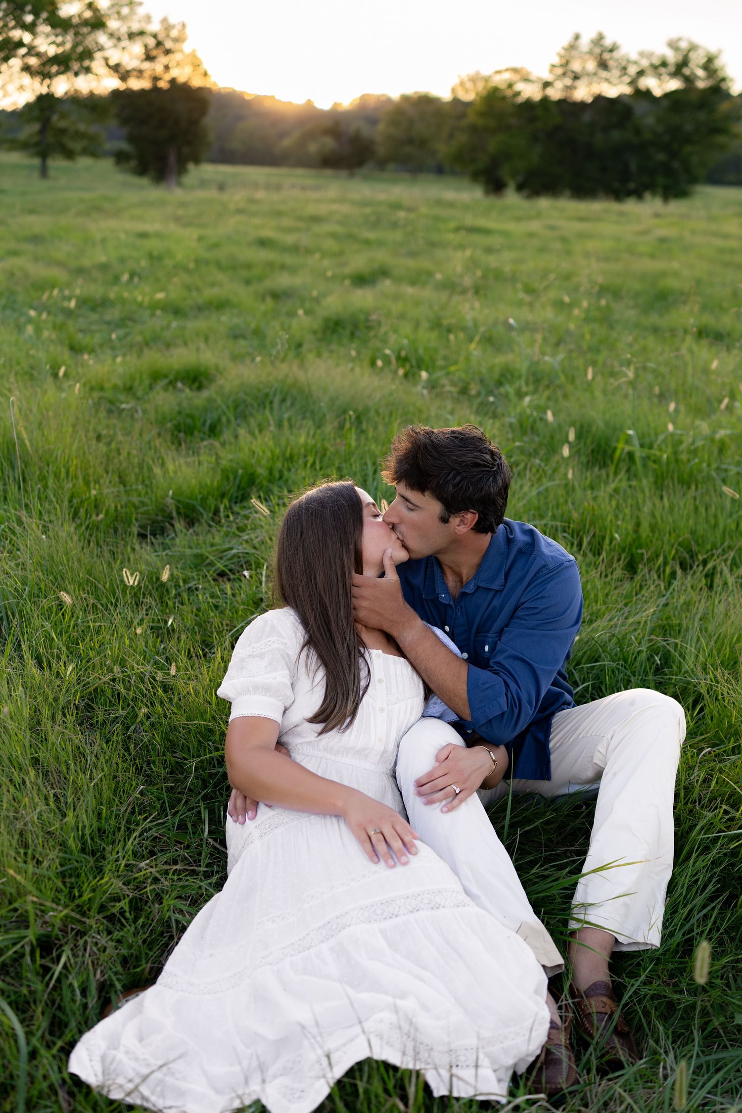 Couple sharing a romantic kiss while sitting in a lush green meadow at golden hour sunset.