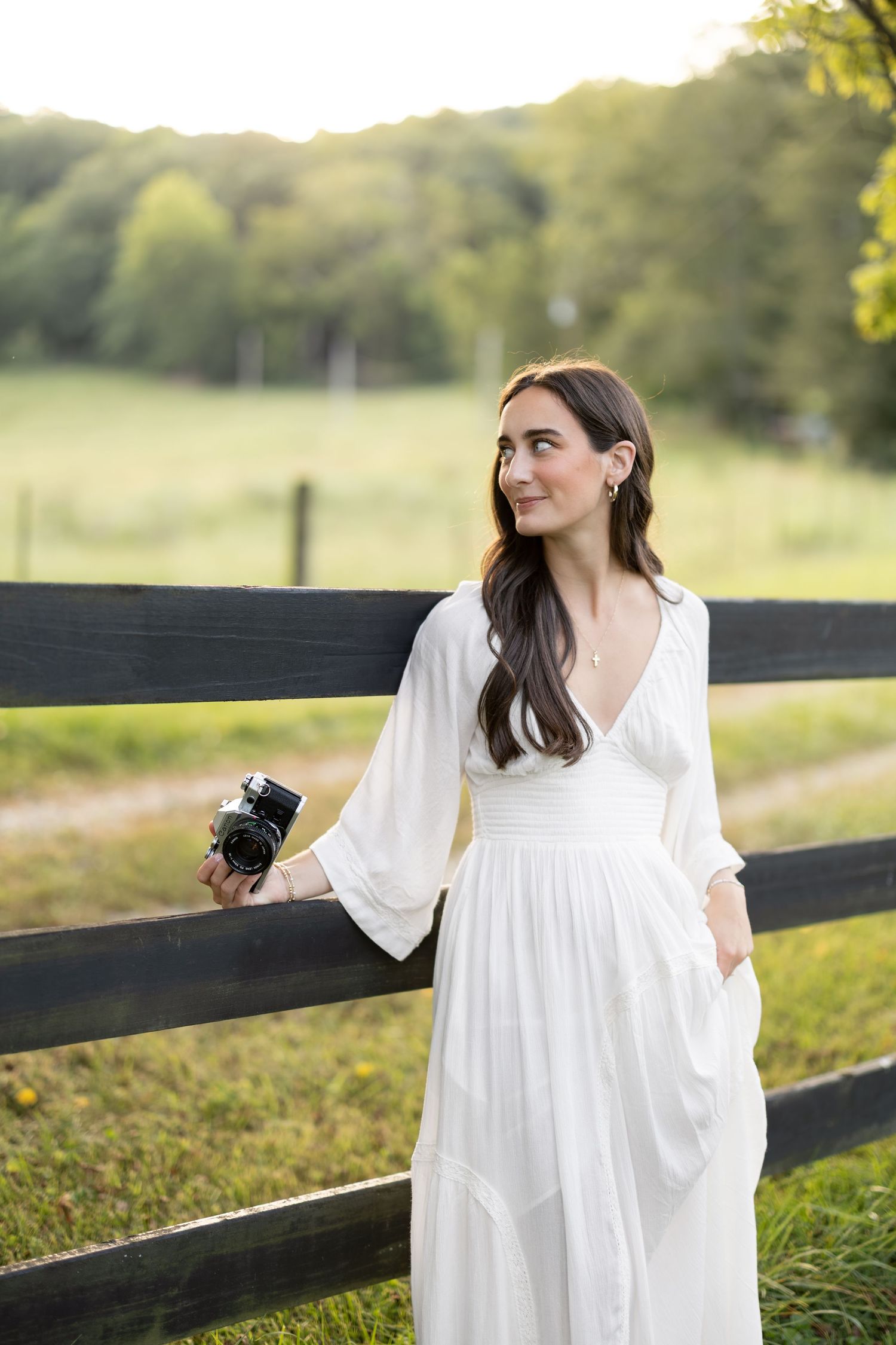 Woman in white dress holding camera leaning on wooden fence in green countryside setting.