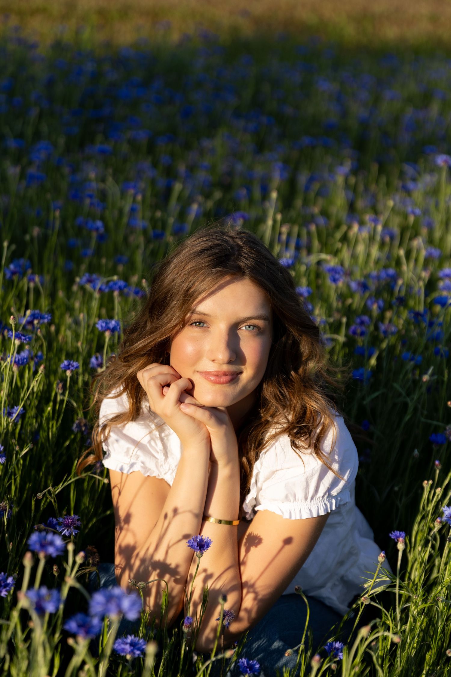 A smiling young woman sits among blue wildflowers in a sunlit field at golden hour.