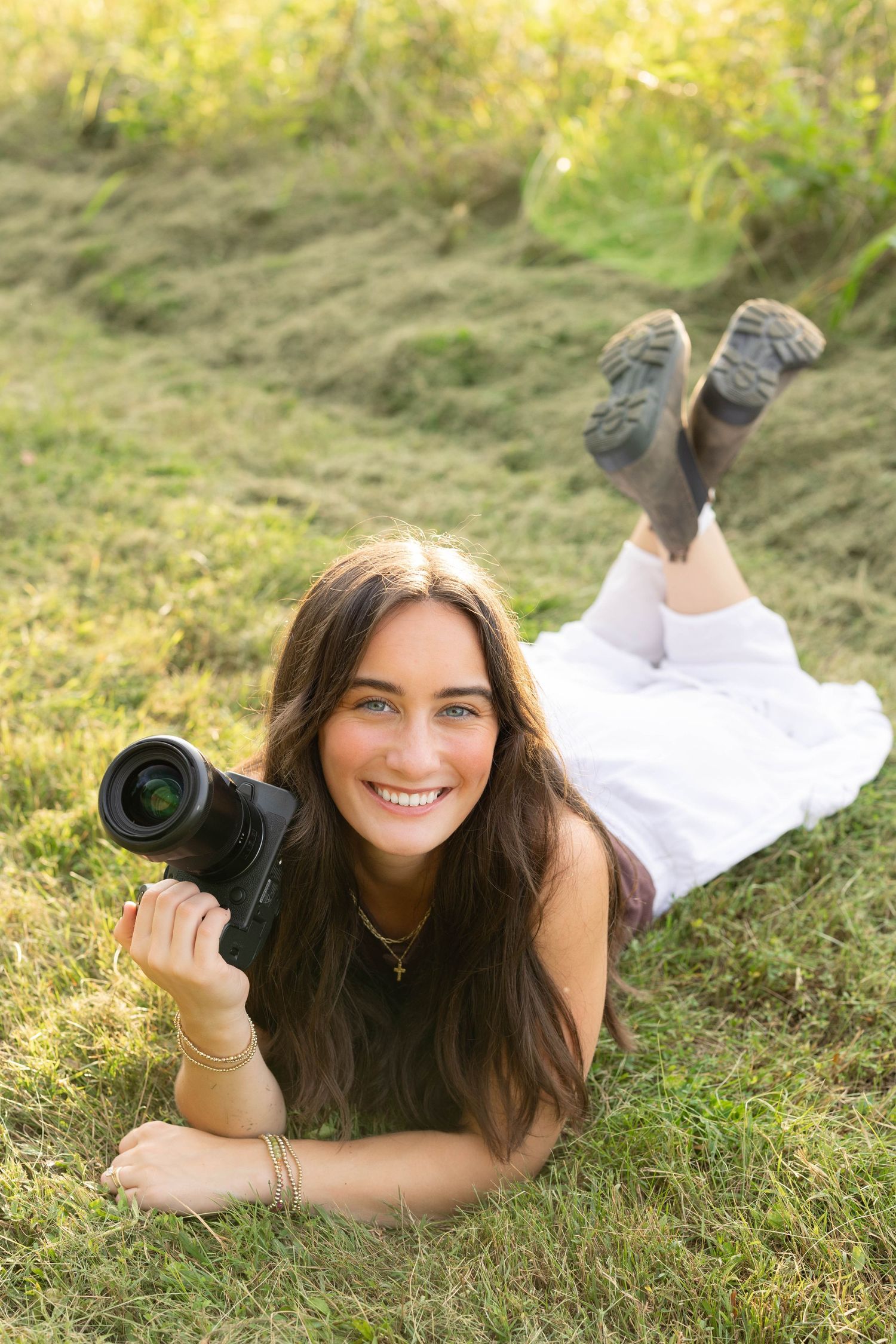 Smiling young woman lying on grass holding DSLR camera in golden afternoon sunlight.