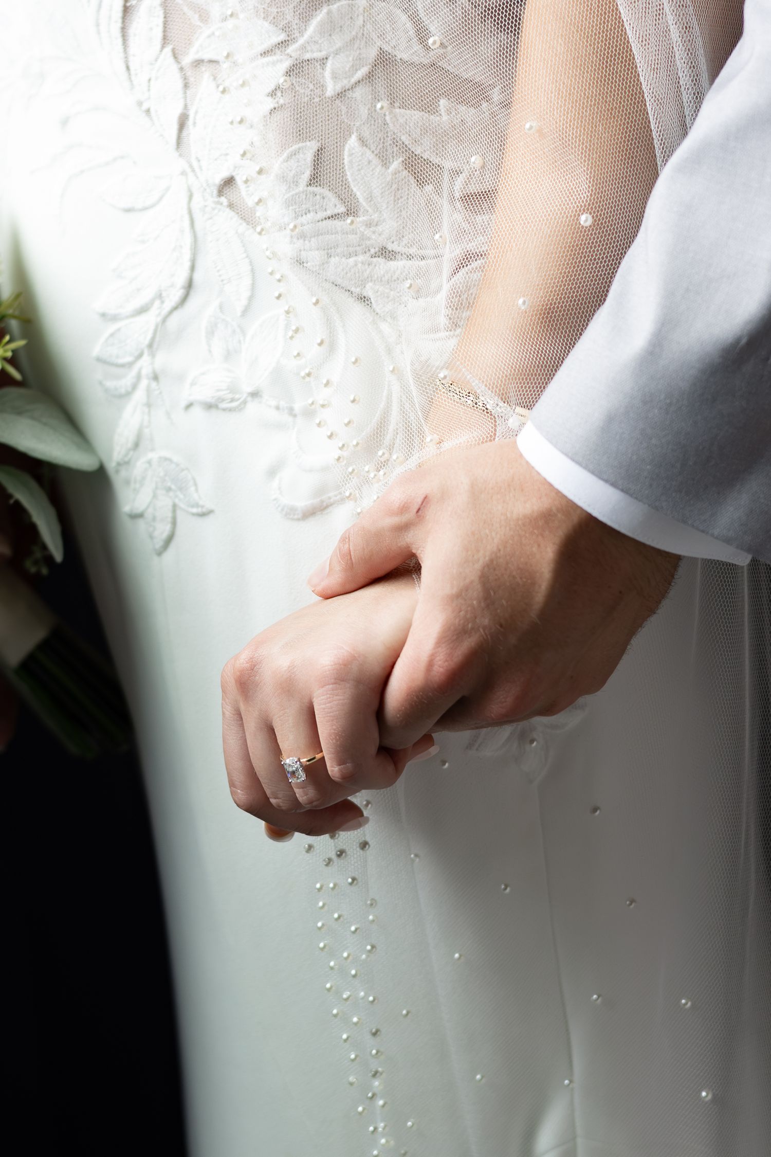 Close-up of a bride and groom holding hands, showing a diamond engagement ring, beaded lace wedding gown and pearl-dotted veil.