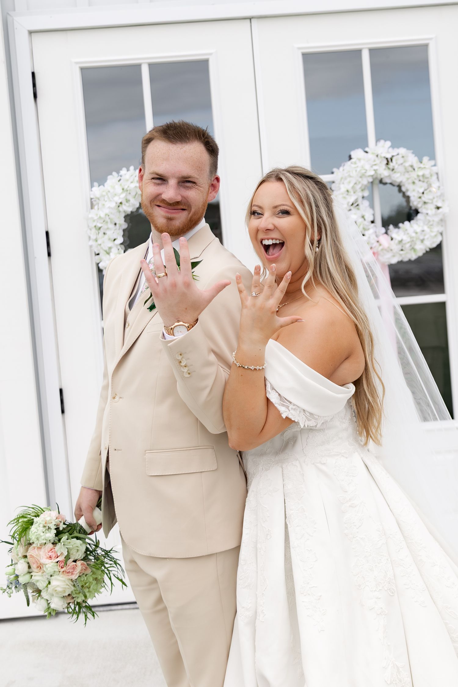 Happy newlyweds showing wedding rings, bride in white gown with veil, groom in beige suit, posing outside white venue.