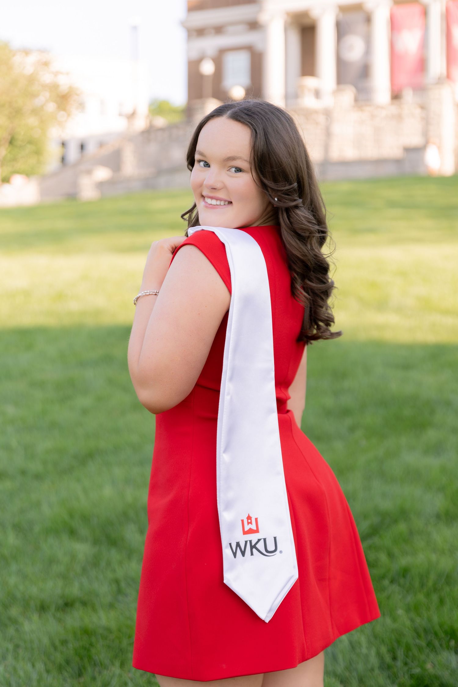 Smiling young woman in red dress with WKU graduation sash poses outdoors on green campus lawn.