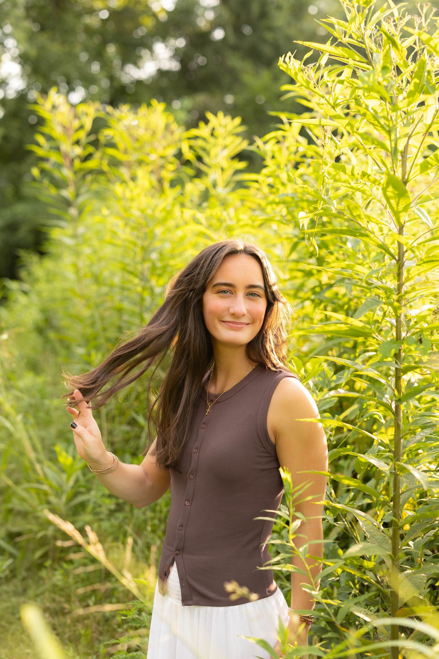 Young woman with long dark hair smiling among tall green plants in a sunlit outdoor setting.