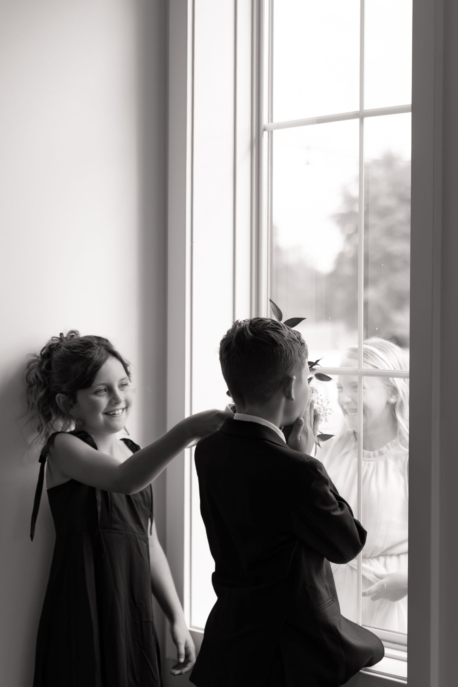 Black and white photo of two children in formal wear looking out a large window, girl smiling at camera.