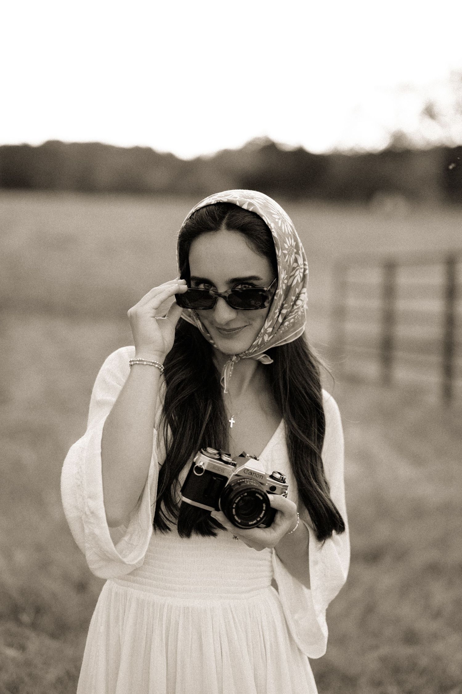 Vintage-style black and white photo of a woman in white dress holding a retro camera, wearing sunglasses and head scarf outdoors.