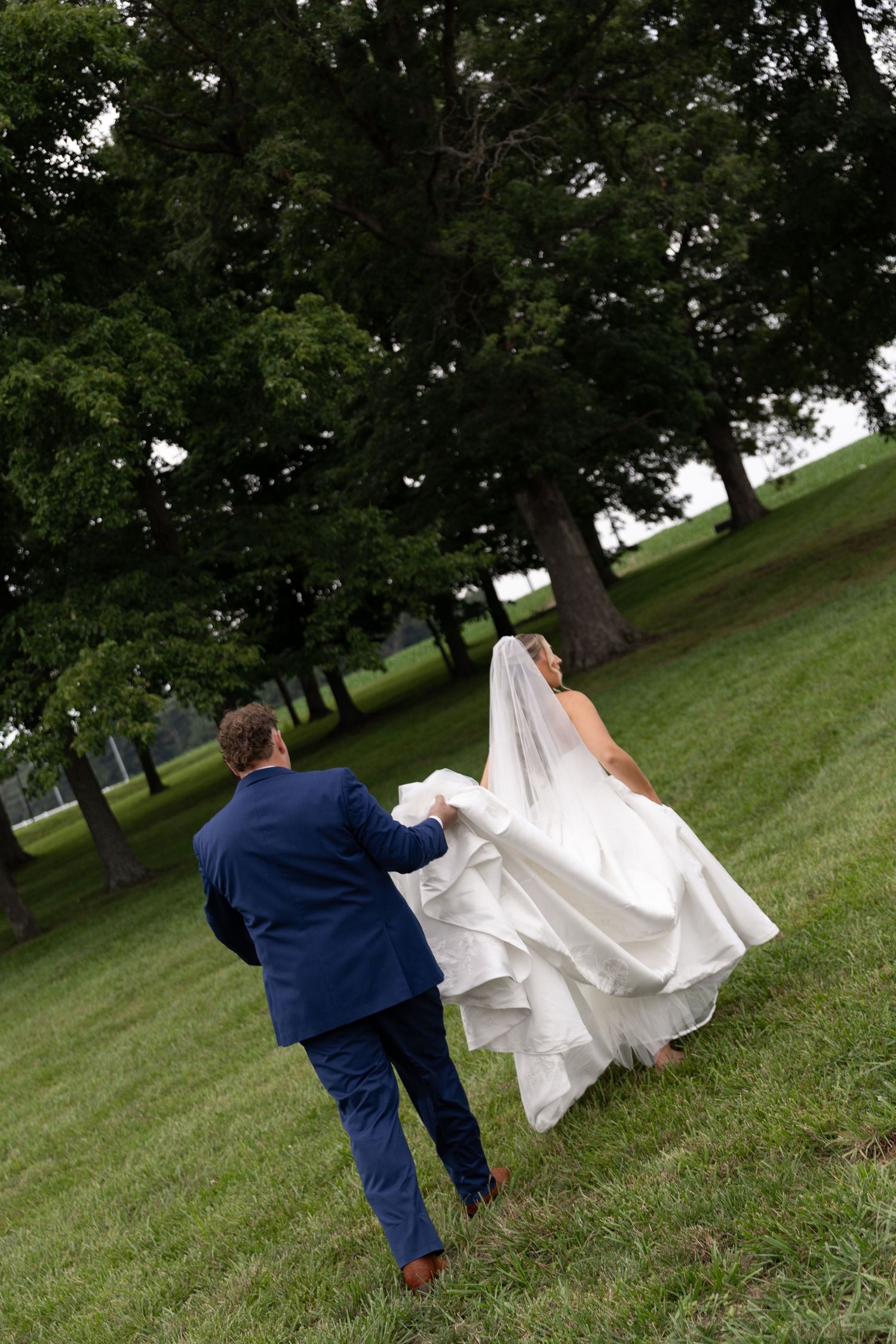 Bride and groom walking hand in hand across green grass park, groom in navy suit holding brides white wedding dress train.