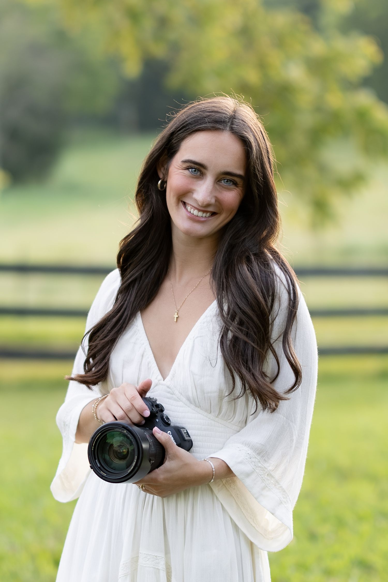 Smiling young woman in white dress holding a DSLR camera outdoors near a wooden fence.
